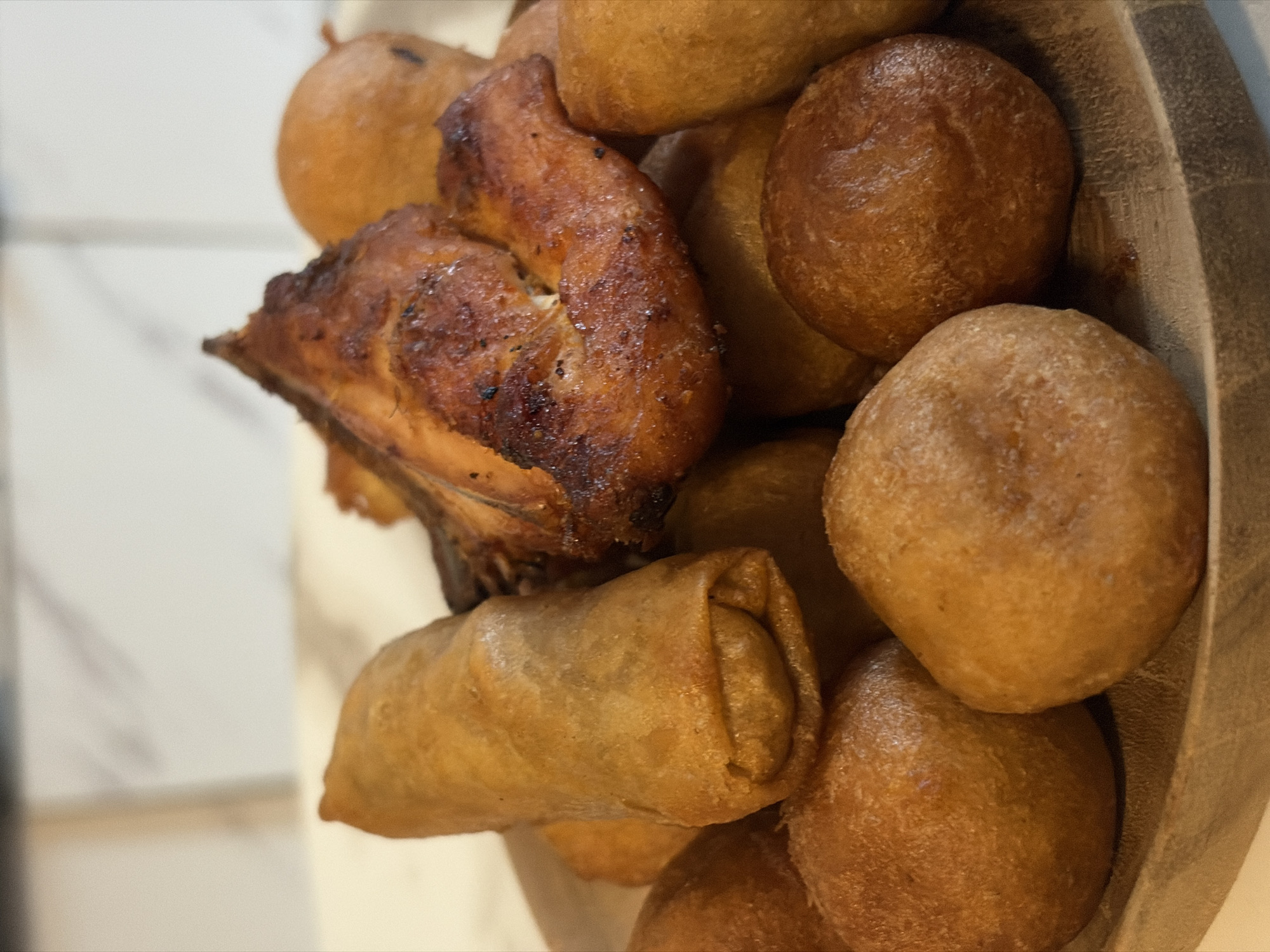 A close-up, top-down shot shows a wooden bowl filled with various foods. The most prominent item is a piece of fried chicken with dark, crispy skin. There is also what appears to be a spring roll. The rest of the bowl is filled with numerous round, golden-brown dough balls. These objects appear to be in a setting, possibly in Igboefon, Nigeria.
