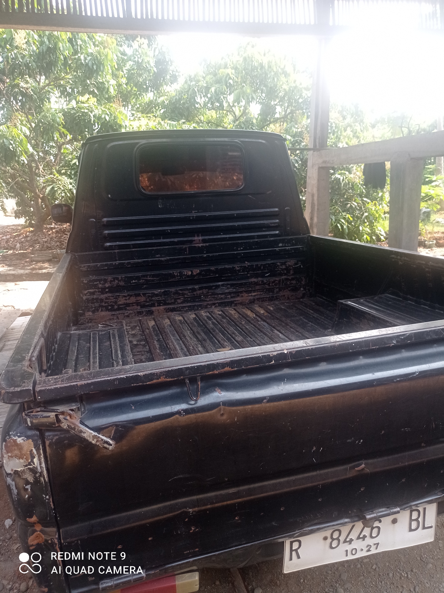 A black pickup truck is parked in an outdoor area with a corrugated metal roof overhead and trees in the background. The truck bed is empty and shows signs of wear and tear, with rust and dirt visible. The license plate is visible and reads "R 8446 BL" with "10.27" below it. The location is identified as Cipawon, Indonesia. The scene appears to be a residential or informal storage area.