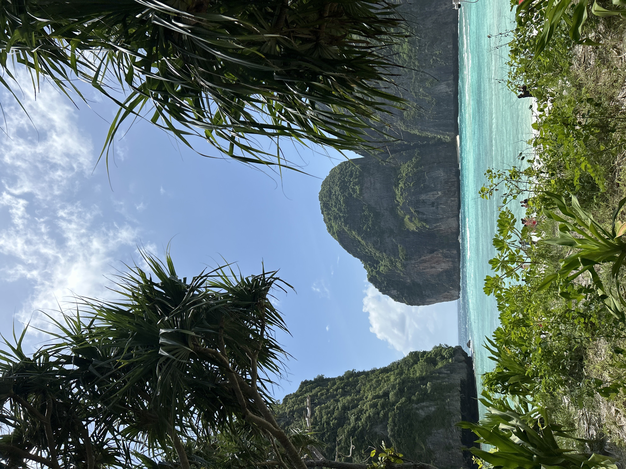 The image depicts a tropical beach scene with lush green foliage in the foreground and turquoise water. In the background, towering limestone cliffs rise dramatically from the sea, a characteristic feature of the Phi Phi Islands, which are accessible from Ao Nang, Thailand. The sky is a vibrant blue with scattered white clouds. The water is clear and calm, with gentle waves lapping the shore. There are a few people visible on the beach, some wading in the water and others relaxing. The overall impression is one of a serene and picturesque paradise.