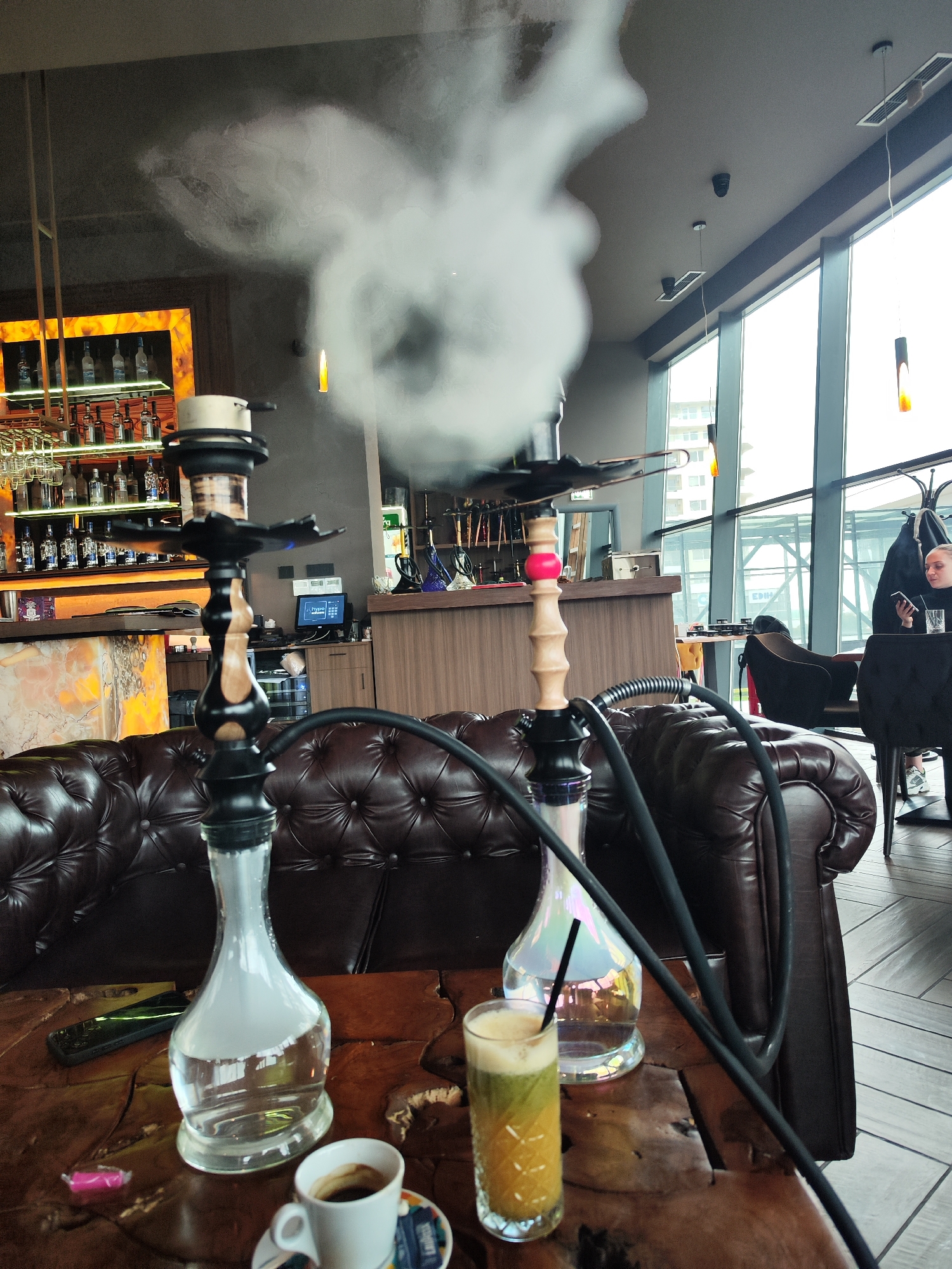 This image presents an indoor scene within what appears to be a modern hookah lounge, bar, or cafe. Two prominent hookahs (shishas) dominate the foreground, resting on a textured wooden table. One hookah has a clear glass base, and the other an iridescent one, both featuring black hoses. A thick plume of white smoke rises from the hookah closer to the viewer, indicating ongoing use.
On the table, alongside the hookahs, are a white espresso cup on a saucer with "LAVAZZA" visible, a tall glass containing a layered green and yellow drink with a straw, a black smartphone, and a pink candy wrapper. Comfortable, tufted brown leather couches provide seating in the foreground.
In the mid-ground, a bar area is visible, stocked with numerous bottles of alcohol, some illuminated from behind. A monitor screen behind the bar displays the word "hype." Further back, large, floor-to-ceiling windows reveal an urban environment with buildings outside, suggesting it is daytime or early evening. The ceiling is gray, featuring modern pendant lights and air vents.
A sole woman is seated at a dark table by the window in the background, focused on her smartphone. She appears relaxed, and a coat rack with a jacket is positioned near her. The overall atmosphere suggests a leisurely and social setting in a stylish establishment, likely in Burgas, Bulgaria.