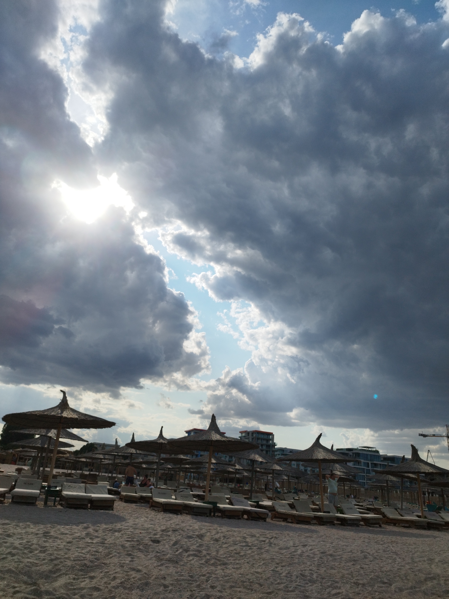 The image depicts a beach scene in Constanța, Romania, on a cloudy day with the sun breaking through the clouds, creating bright shafts of light. The foreground is a sandy beach with rows of lounge chairs and straw umbrellas. In the mid-ground, several people are visible, some lounging on chairs and others standing. Further back, modern buildings line the coast, with a construction crane visible on the right side of the image. The sky is dominated by dramatic, dark clouds, interspersed with patches of blue sky.