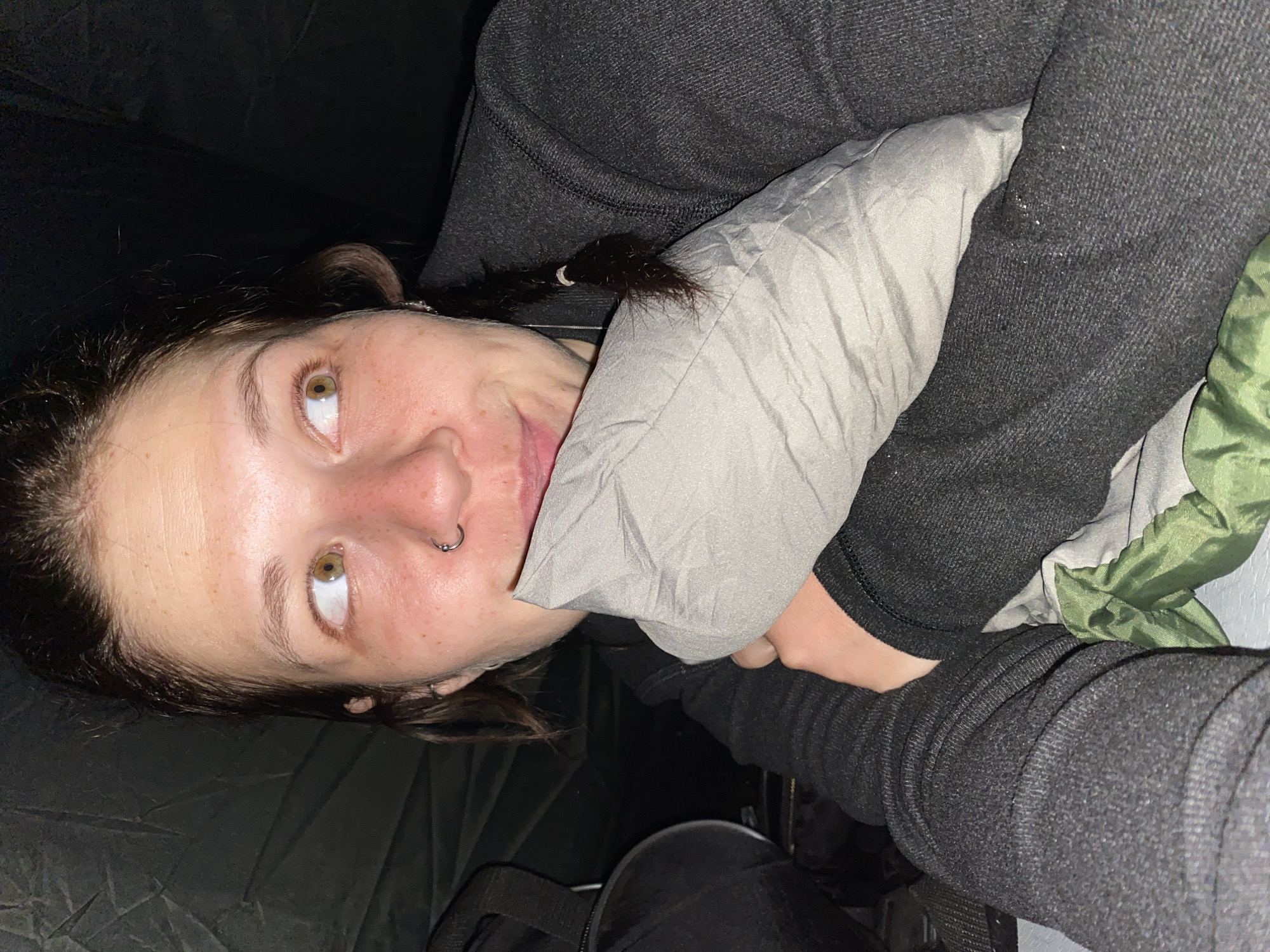 The image shows a close-up, low-angle shot of a young Caucasian woman lying down, holding a gray pillow. She has light skin with freckles and is looking to her right. Her hair is dark brown and pulled back in a braid on the right side, with some wisps framing her face. She is wearing a dark gray hooded jacket. A nose ring is visible on her septum. The background is dark and appears to be the interior of a tent, with a dark green sleeping bag or fabric visible at the bottom. A black bag is partially visible to the left of the frame. The lighting is dim, suggesting it is nighttime or a dark indoor environment. The scene is likely in Абзаново, Russia, given the context.