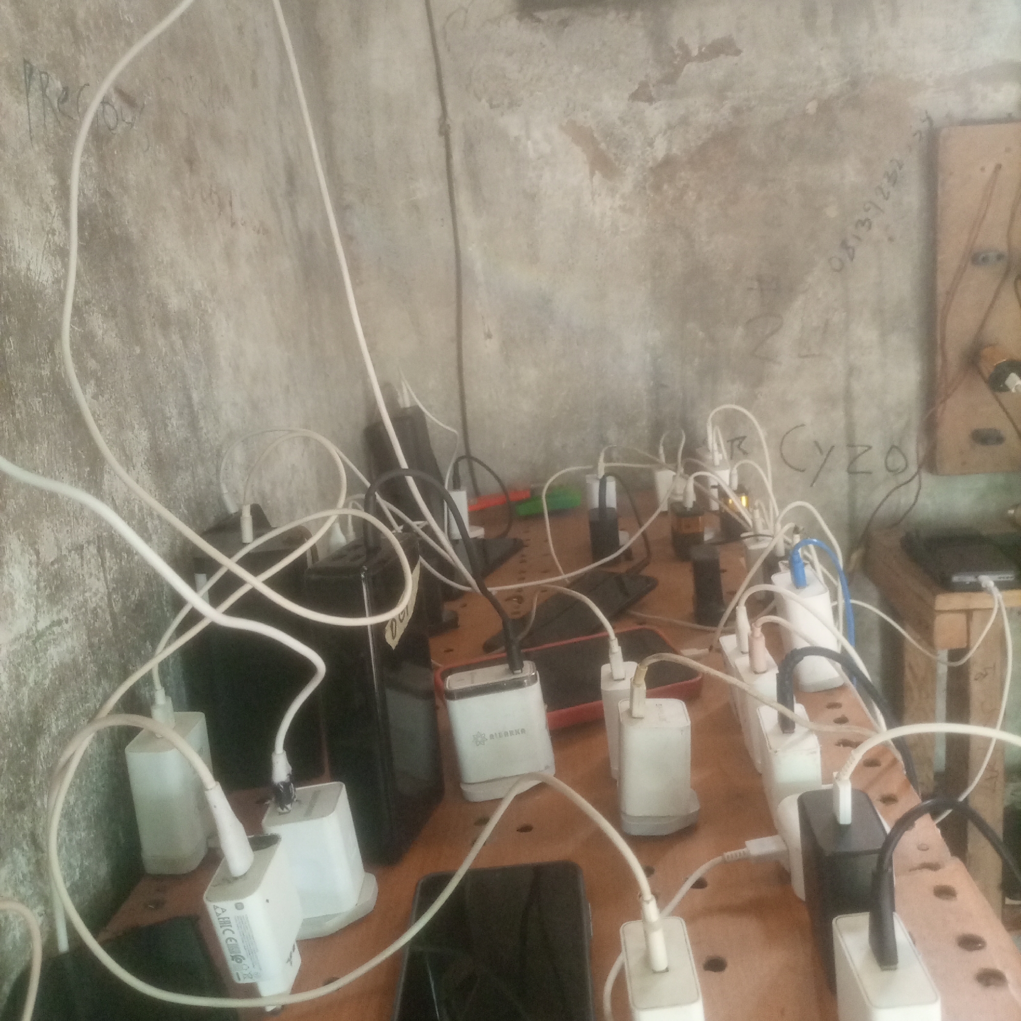 The image shows a cluttered surface, likely a table or shelf, with numerous cell phone chargers and cables. Several white and black phone chargers are plugged into power outlets, with white and some black cables connecting them to various phones and other devices, most of which are not clearly visible. Some phones appear to be lying flat on the surface, plugged in. The background is a textured, concrete-like wall with some faint writing on it. The overall impression is of a makeshift charging station. There is no visible indication of the location being in the city of Unknown, Unknown.