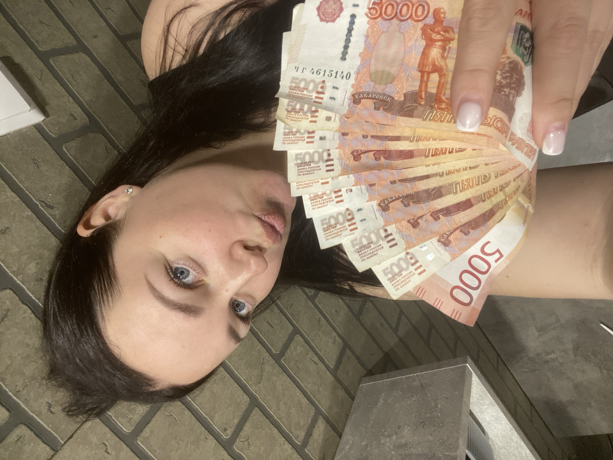 The image captures a woman with dark hair and light blue eyes, looking directly at the camera. Her head is tilted, and she is holding a fan of multiple 5000 Russian ruble banknotes in her right hand. Her nails are neatly manicured in a French style. A small stud earring is visible in her left ear. The background consists of a dark, brick-patterned surface, suggesting an indoor setting or a patio.
The activity depicted is the woman posing with a substantial amount of cash. Her expression is neutral to slightly serious.
Notable details include the crisp appearance of the banknotes, indicating they are relatively new. The lighting is somewhat dim, suggesting an indoor environment or evening time. The Russian currency clearly places the location in Russia, aligning with the provided context of Sochi.
Visible text on the banknotes includes the denomination "5000" prominently, along with "ПЯТЬ ТЫСЯЧ РУБЛЕЙ" (Five Thousand Rubles). A serial number "ЧГ 4615140" is visible on one note. The city "ХАБАРОВСК" (Khabarovsk) is also discernible, referring to one of the cities depicted on the 5000 ruble note. Additionally, the security warning "ПОДДЕЛКА БИЛЕТОВ БАНКА РОССИИ ПРЕСЛЕДУЕТСЯ ПО ЗАКОНУ" (Counterfeiting banknotes of the Bank of Russia is punishable by law) can be seen on several notes.
