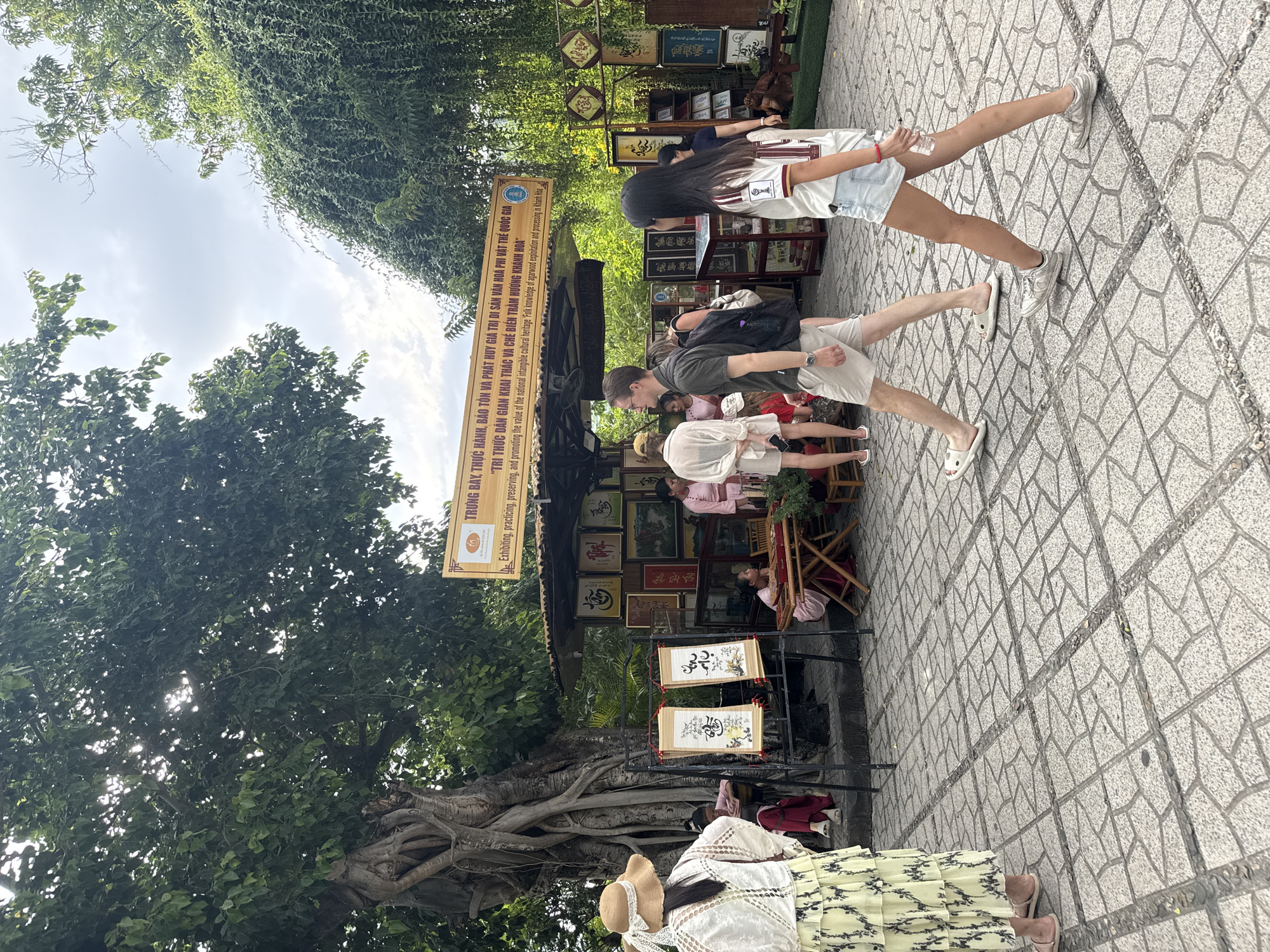 The image depicts a scene at a market stall in Nha Trang, Vietnam, likely within a cultural or historical site. In the foreground, a woman with a straw hat and a light-colored patterned skirt is seen from behind, facing a display of calligraphy scrolls. Two people, a man in a grey t-shirt and khaki shorts and a woman in a white t-shirt and denim shorts, walk past the stall from left to right. Behind the people and the stall, a large, ancient tree with exposed roots is visible. The stall itself is covered by a corrugated roof and displays various framed artworks, predominantly calligraphy and paintings. Several other people are present, some browsing the art, and one appearing to be a vendor sitting at a low wooden table. The ground is paved with decorative grey tiles. A yellow banner with Vietnamese text and English translation hangs above the stall, indicating that it is part of an exhibition and promotion of the value of intangible cultural heritage. The overall atmosphere suggests a tourist-oriented cultural attraction.