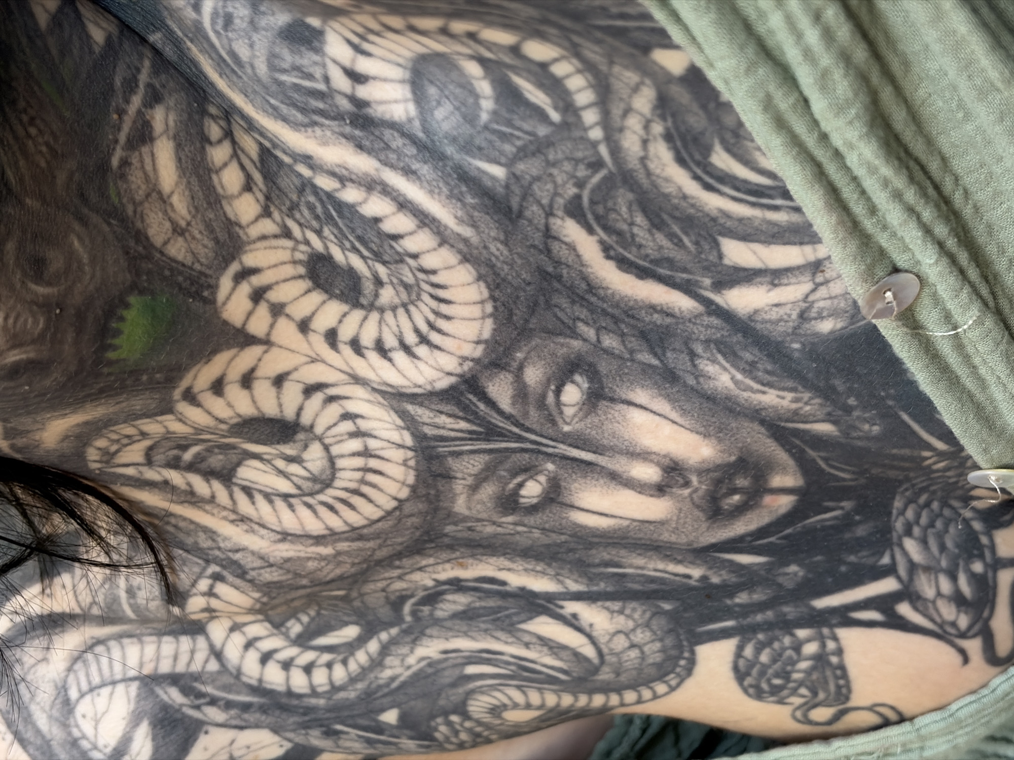 The image depicts a close-up view of a tattoo on human skin, featuring a Medusa-like figure with snakes as hair. The tattoo is rendered in black and grey tones, with a high level of detail in the depiction of the snakes and the face. A portion of a light green garment with two buttons is visible on the right side of the image. The presence of the tattoo and clothing are the most identifiable elements. There is no information to suggest that this image was taken in the city of Chaikovsky, Russia.