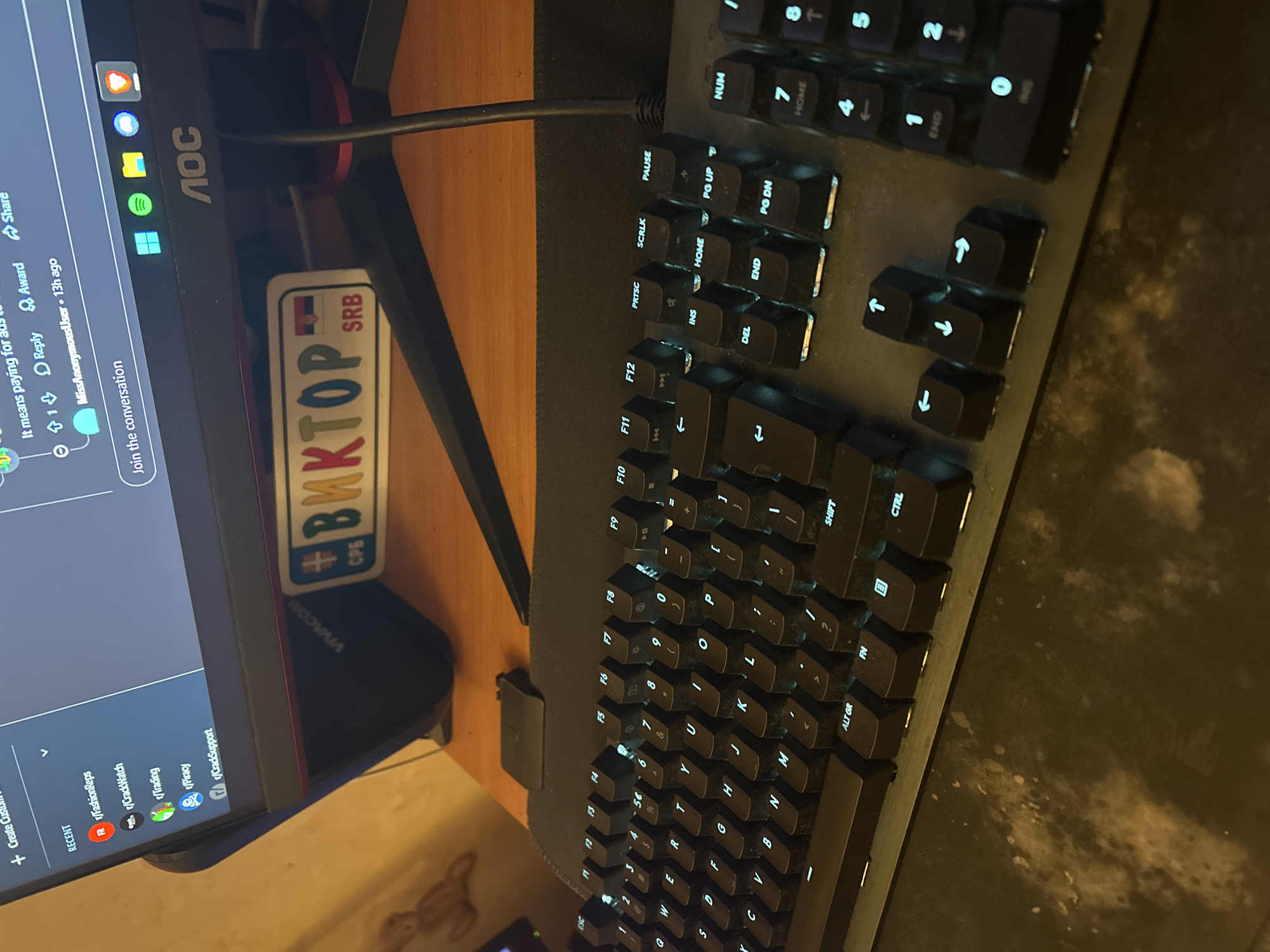 The image captures a close-up, high-angle view of a computer workstation setup, likely indoors. The main subjects are a backlit keyboard and a monitor screen, positioned on a dark mouse pad that covers a wooden desk surface.
The monitor, branded "AOC", displays a social media interface resembling Reddit. Visible text on the screen includes a post title "It means paying for a...", a comment from "MissAnonymousUser • 13h ago", and options like "Reply", "Award", and "Share". A "RECENT" section lists several subreddits: "r/FashionReps", "r/CrackWatch", "r/Trading", "r/Piracy", and "r/CradSupport". The monitor stand features the "VIVACOM" logo, identifying a Bulgarian telecommunications company. Attached to the monitor stand is a sticker with a Serbian flag and the text "ВМКTOP" (BMKTOP in Cyrillic) and "SRB", indicating a connection to Serbia.
The keyboard, a dark model with light blue or white backlighting, shows various keys, including QWERTY letter keys, number keys (1-0), function keys (F1-F12), and special keys such as PRTSC, SCRLK, PAUSE, INS, HOME, PG UP, DEL, END, PG DN, NUM, SHIFT, CTRL, ALT GR, FN, ESC, and arrow keys. The keyboard appears to be a mechanical type due to the raised keycaps.
The scene depicts a typical personal computer environment in Sofia, Bulgaria, evidenced by the "VIVACOM" branding. The Serbian sticker suggests a possible personal or cultural connection to Serbia. The time of day is not discernible, as the setting is illuminated by indoor lighting. No specific activity beyond browsing is clearly indicated, and no emotions are discernible from the inanimate objects.