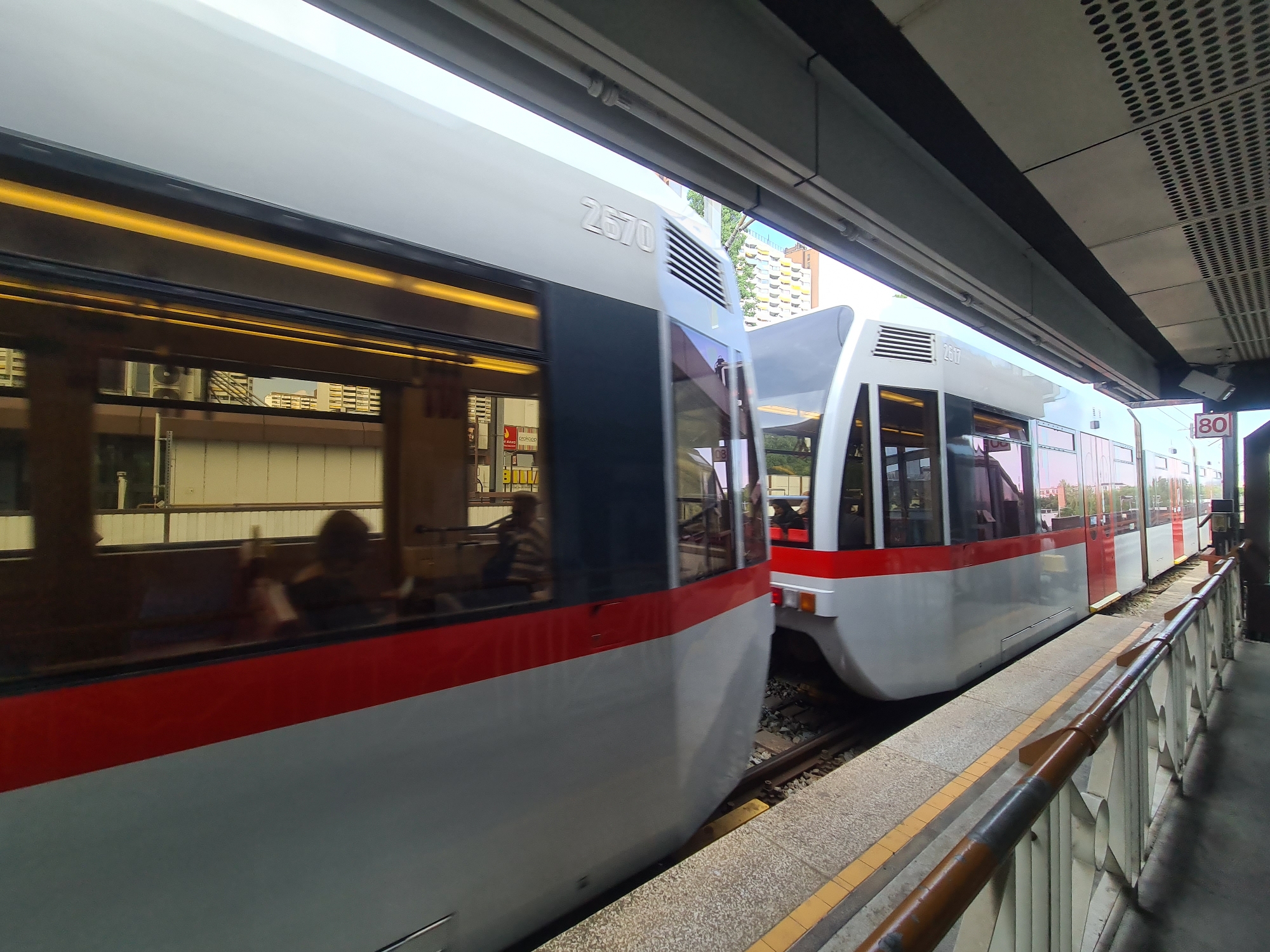 A low-angle, slightly motion-blurred exterior shot captures two white and red trams, numbered 2570 and 2517 respectively, stopped at a station. The tram on the left, closer to the viewer, is largely in focus, with its side featuring large windows reflecting buildings. The tram on the right is partially obscured by the first and appears to be moving away. The station platform, made of concrete with yellow and grey markings, is visible in the foreground. A brown metal railing lines the edge of the platform. Overhead, a modern, perforated ceiling structure is supported by grey beams. In the background, buildings with visible air conditioning units and apartment facades are seen under a bright sky. A red sign with a white "80" is mounted on a pole to the right of the tram. The scene is set at the Handelskai U-Bahn station in Vienna, Austria, a transportation hub connecting the U6 and U2 lines.