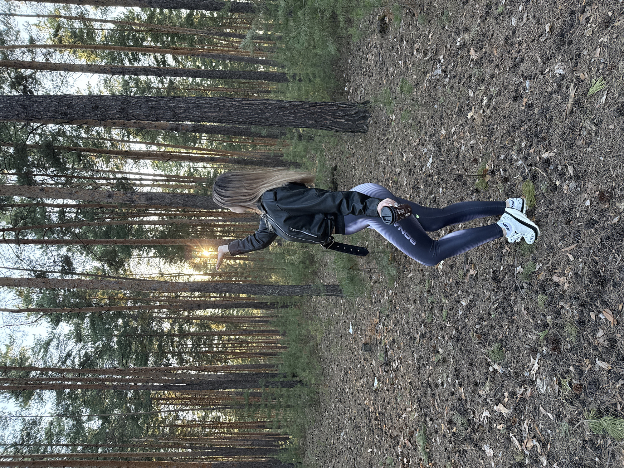 The image depicts a person, appearing to be a young woman, standing in a sun-drenched pine forest. She is viewed from behind, facing away from the camera, with her long, light-colored hair cascading down her back. She is dressed in a black leather jacket, form-fitting dark grey or black leggings, and white athletic shoes with dark accents. A black belt is visible around her waist.
Her left arm is extended towards the left side of the frame, with her palm open, seemingly reaching for or interacting with the bright, golden sun visible through the trees. In her right hand, she holds a dark object, possibly a camera lens or a small container. The text "BONA" is clearly visible in white letters running vertically down the outer thigh of her left legging.
The setting is a dense pine forest, characterized by numerous tall, slender trees with rough bark. The ground is covered with a thick layer of dry pine needles, fallen leaves, and small twigs, indicating a natural, unpaved environment. The strong, low-angle golden sunlight breaking through the trees suggests either early morning or late afternoon. The sky is partially visible through the canopy, appearing bright. The overall impression is one of a peaceful, natural outdoor scene.