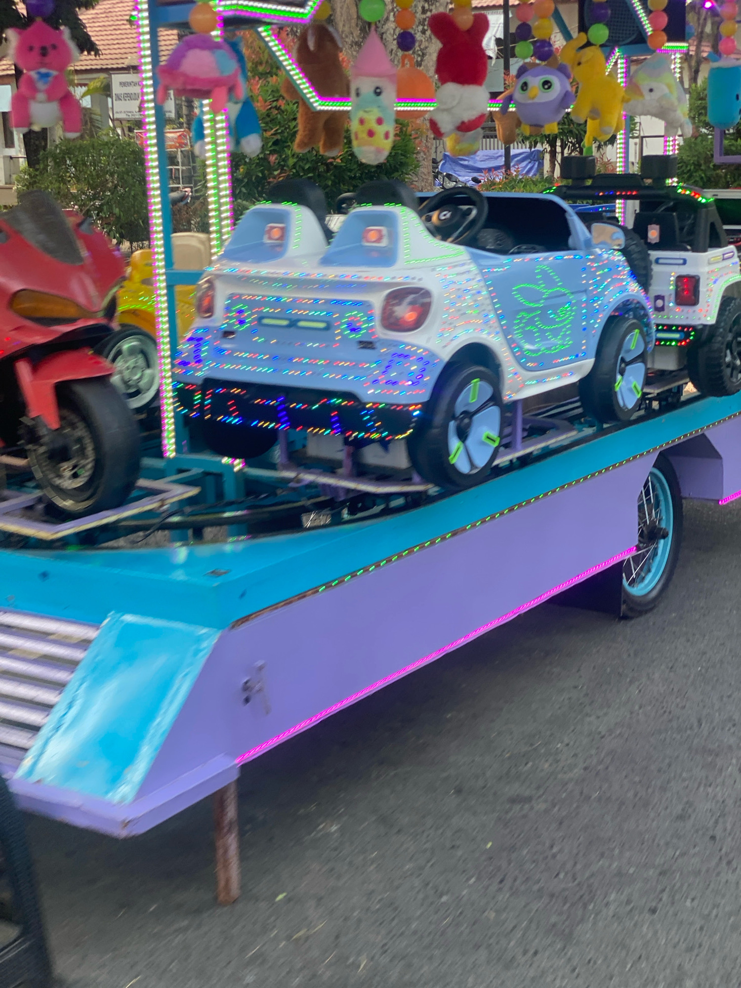 The image shows a close-up, low-angle shot of a brightly lit amusement ride. The ride consists of a trailer with a purple and turquoise platform adorned with pink and green LED lights. On the platform are several child-sized electric cars, including a red motorcycle and two stylized blue and white cars decorated with numerous colorful LED lights. The cars are positioned on a raised track. In the background, a building with trees and a white fence is visible, suggesting an outdoor setting. The blurred motion indicates the ride is either in operation or has recently been in motion. The presence of amusement rides and the surrounding environment are consistent with a location in the city of Banjarbaru, Indonesia, likely an entertainment area or fairground.
