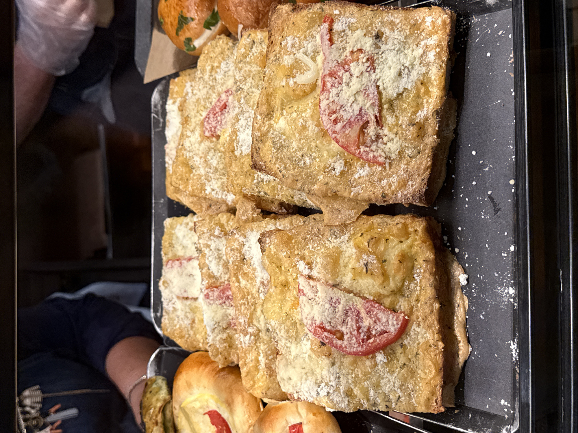 The image shows a close-up, eye-level view of a tray of baked goods, possibly in a bakery or food display, in Kadıköy, Turkey. The tray, made of a dark, matte material, is filled with rectangular slices of what appears to be focaccia or a similar flatbread, topped with grated cheese and pieces of tomato. The baked goods are golden brown and dusted with a light coating of white powder, possibly flour or more cheese. In the background, partially visible, are hands of two individuals. One hand, on the left, is wearing a bracelet and seems to be reaching towards the baked goods. The other hand, on the right, is covered with a clear plastic glove, suggesting food handling. The lighting is dim, with some light sources from above, creating shadows and highlighting the texture of the food. The overall impression is one of fresh, ready-to-eat baked goods presented for sale or consumption.