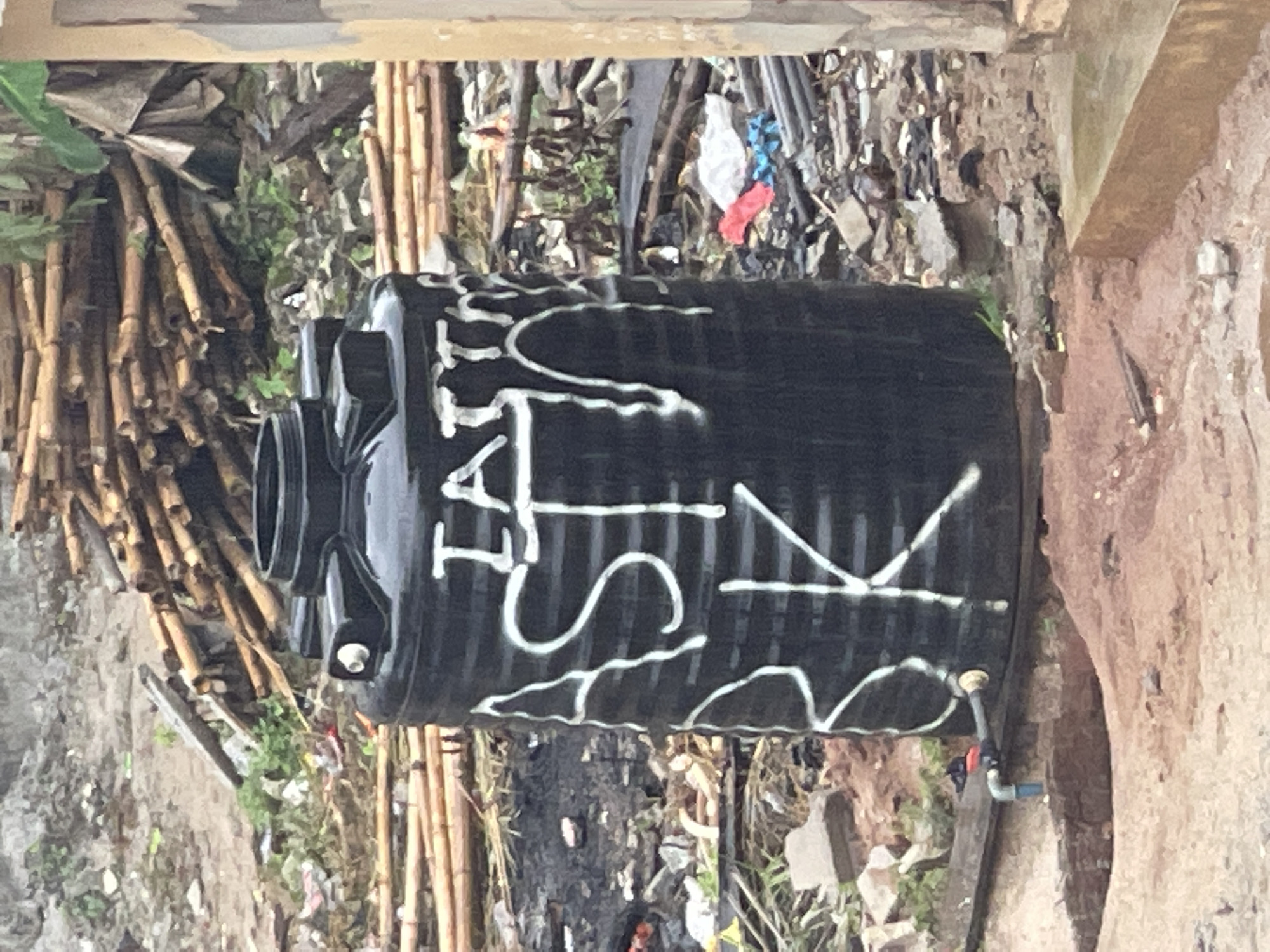 A black plastic water storage tank rests on the ground amidst debris in an outdoor setting, likely in Abraka, Nigeria. White spray paint decorates the tank with the lettering "LAT SIK". The surrounding environment features bamboo poles, discarded materials, and dirt. The tank is positioned near a weathered concrete or earthen wall structure. A small blue and red valve is connected to the tank.