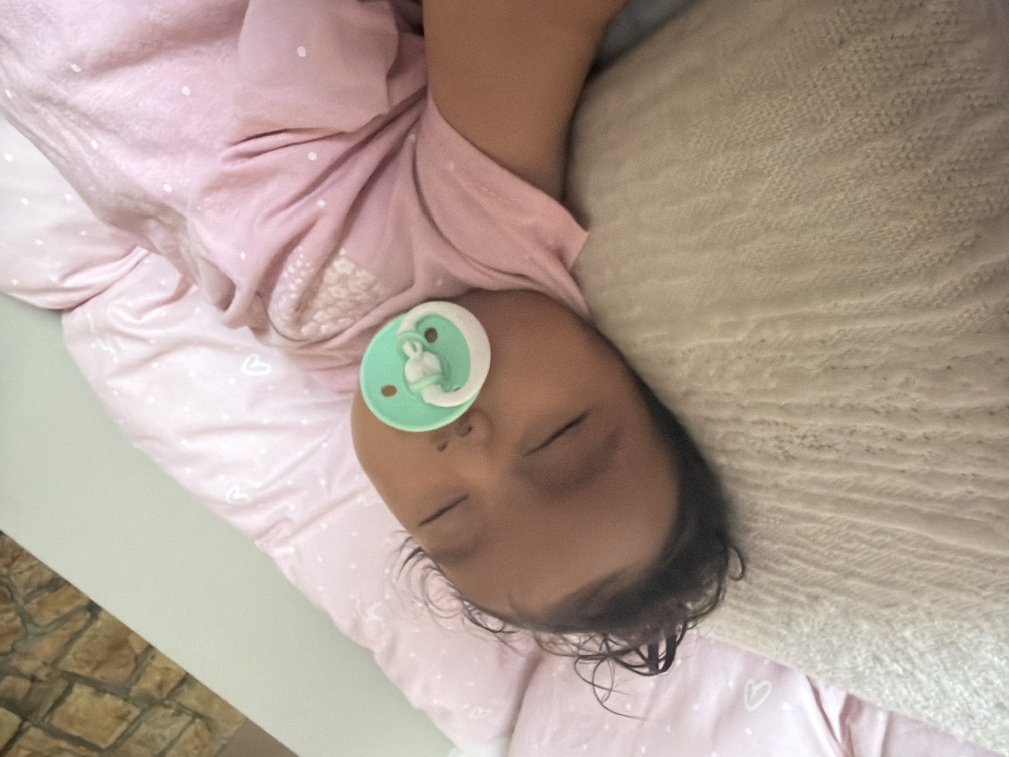 A close-up, overhead shot shows a baby sleeping with a pacifier in its mouth. The baby is lying on a pink blanket with white polka dots, and a light-colored knitted blanket is visible beneath its head. The background shows a textured stone wall. The scene is indoors.