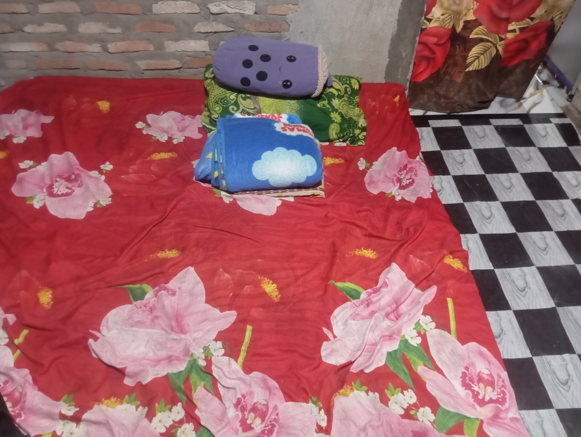 A red floral bedspread is spread over a mattress. On the bedspread, there is a folded blue blanket with a cloud pattern, a green pillow with a paisley pattern, and a purple pillow with black dots resembling a face. The bed is against a brick wall. To the right of the bed is a floor with a black and white checkered pattern. A brown and red floral curtain hangs to the right of the bed. The scene appears to be indoors in a room.