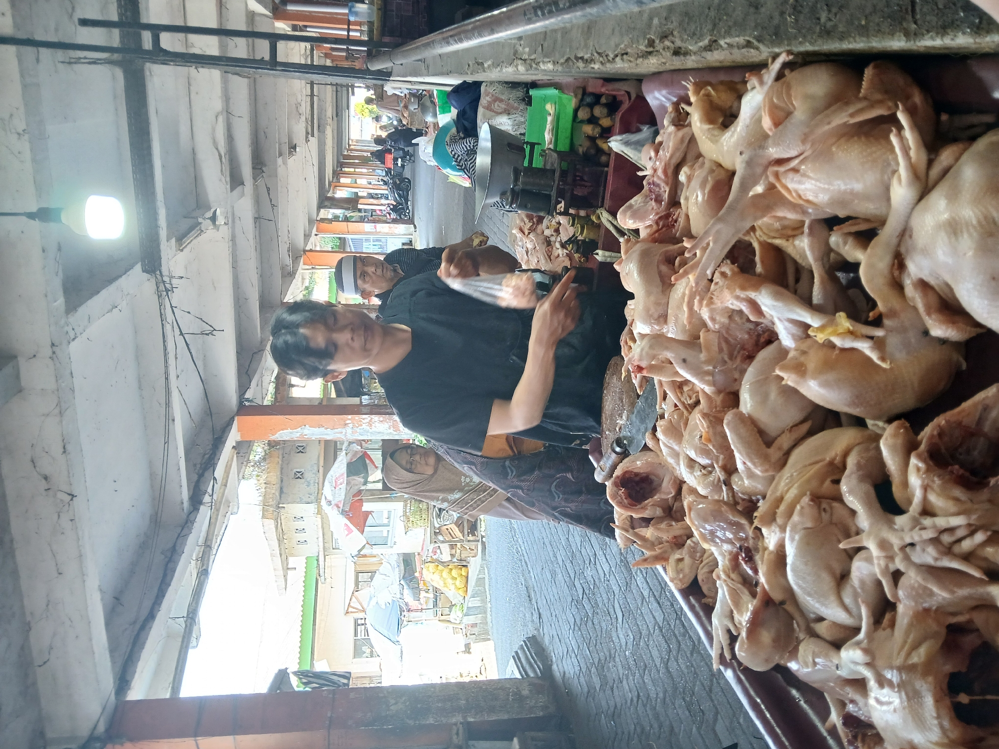 This image depicts a scene at a wet market, likely in Kupang, Indonesia. In the foreground, a vendor, a person with short dark hair wearing a black t-shirt, is handling a plastic bag containing poultry. They are standing behind a large tray filled with whole, uncooked chickens. To the left of the vendor, a woman wearing a hijab and glasses is partially visible. Behind them, another person, a man wearing a white and black checkered cap, is looking towards the camera. The market appears to be an open-air structure with a concrete ceiling supported by pillars. In the background, other stalls, people, and motorcycles are visible, suggesting a bustling marketplace environment. The ground is paved with bricks. The overall lighting suggests it is daytime.