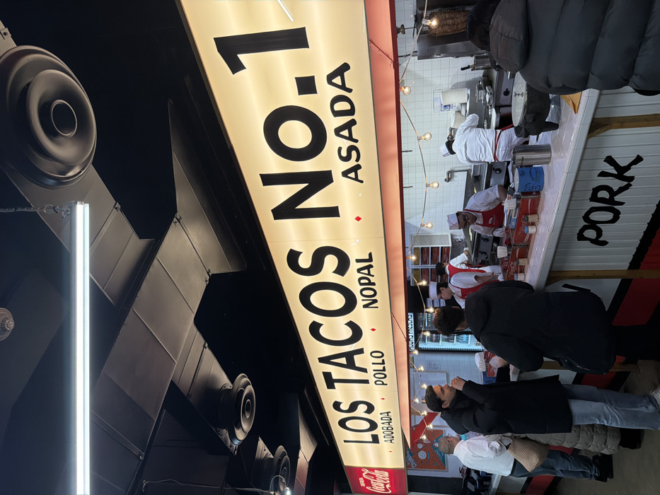 A taco stand sign reads "LOS TACOS NO. 1," listing various meats like Asada, Nopal, Pollo, and Adobada. A Coca-Cola logo is visible beneath the sign. Several customers queue at the counter, where workers in white hats and red aprons prepare food. The kitchen area features tiled walls and visible food preparation equipment. A "PORK" sign is also seen.
