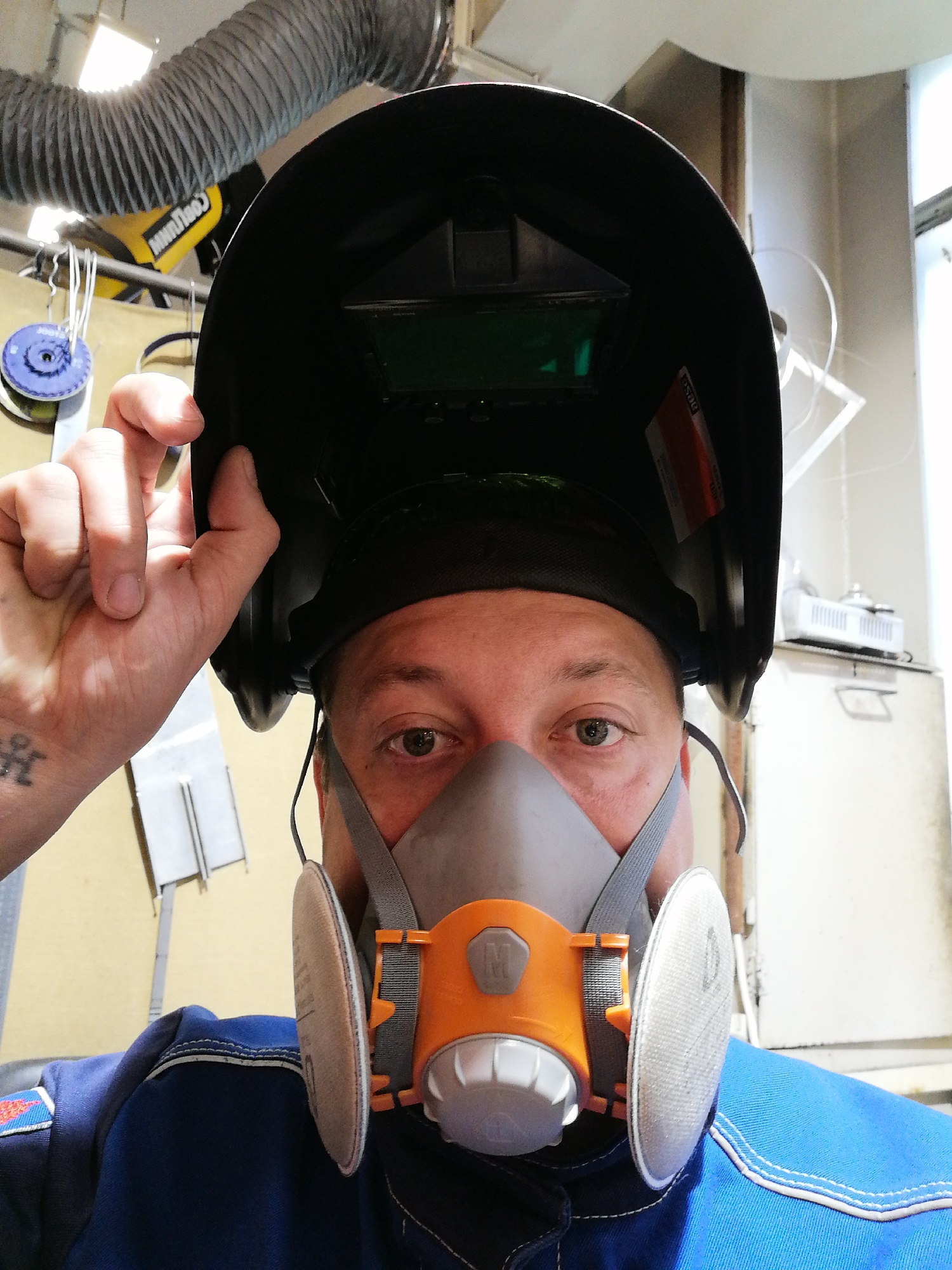 A person wearing a blue jumpsuit and a respirator mask with grey straps and orange accents is holding up a black welding helmet. The person has brown eyes, and a tattoo is visible on their left hand. Above the person, there is a flexible grey duct. The background contains tools and equipment, and the location appears to be a workshop of some kind. The city of Unknown, Unknown is not specified or confirmed.