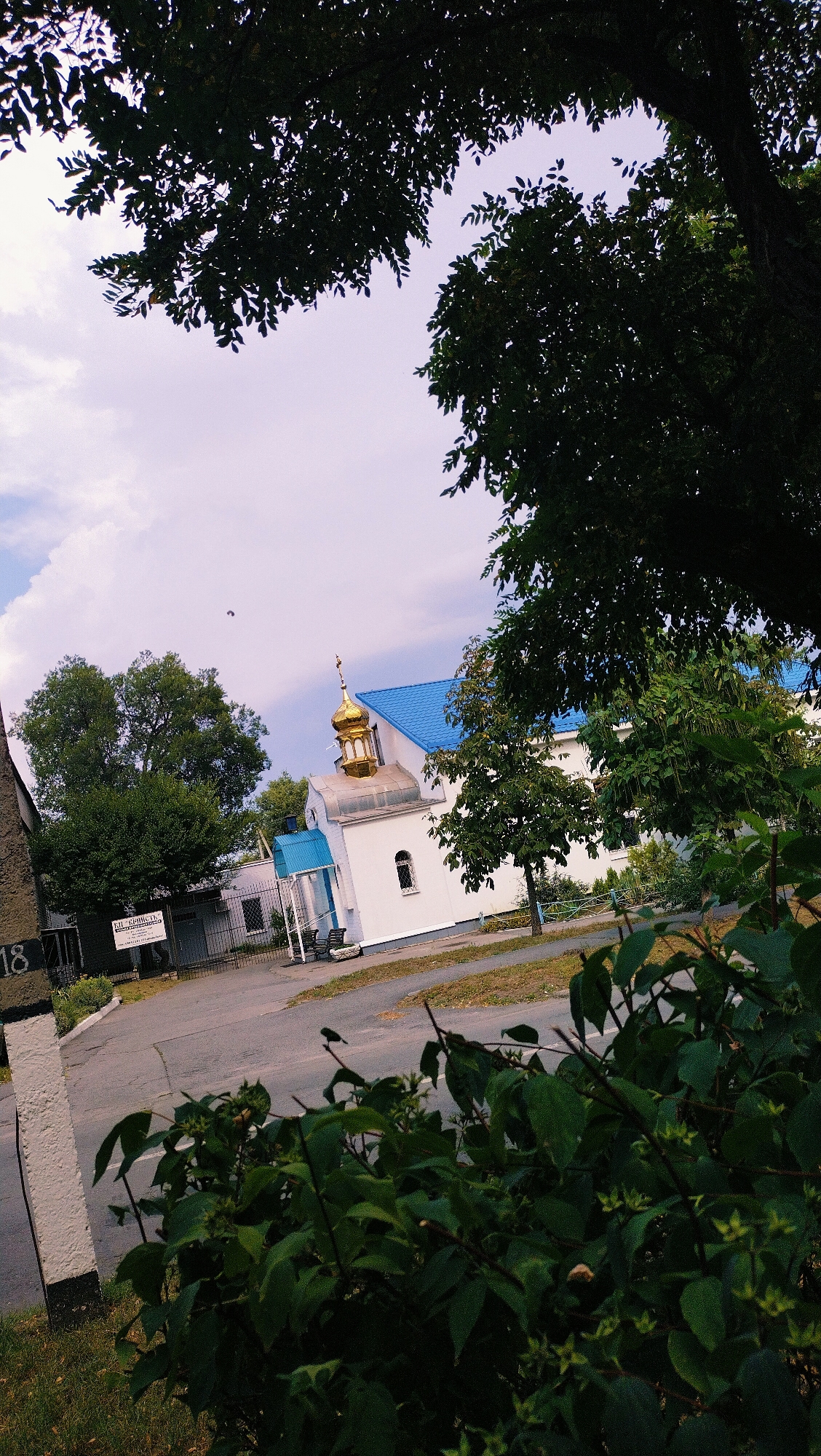 The image depicts a small white building with a golden dome, characteristic of an Orthodox church, with a bright blue roof, situated in the city of Horishni Plavni, Ukraine. The building is surrounded by lush green trees and a cloudy sky, with one tree branch prominently in the foreground, partially obscuring the view. A street or pathway runs in front of the church, and to the left, a portion of another building with the number "18" visible on its side is present. The scene appears to be a quiet, perhaps residential or semi-rural, area within the city.
