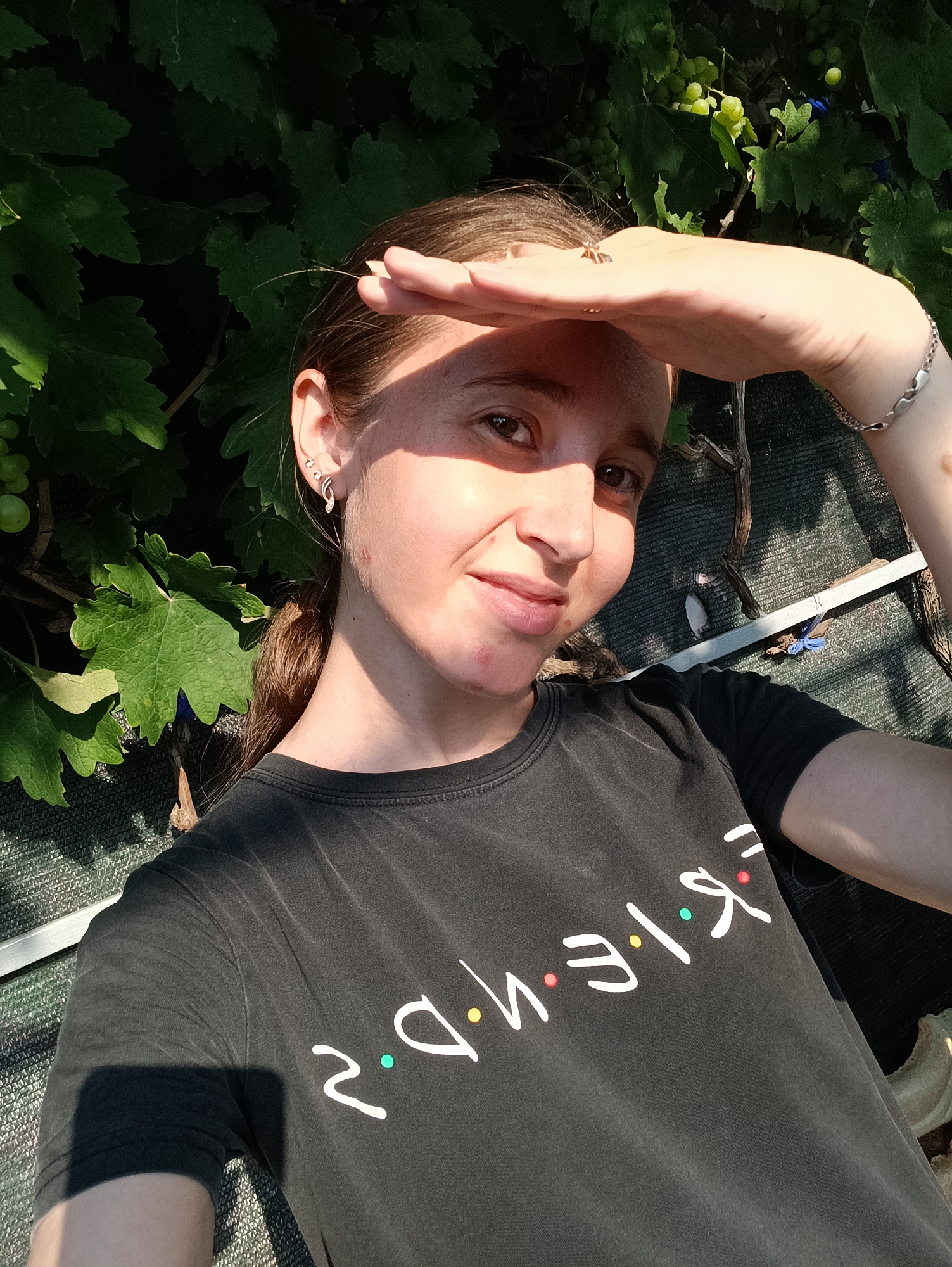 A young woman stands in front of a green leafy vine, likely in Vypasne, Ukraine, on a sunny day. She is wearing a dark t-shirt with the "Friends" logo emblazoned across the front in white lettering with colored dots. The woman is holding her hand up to her forehead, shielding her eyes from the sun as she looks directly at the camera with a slight smile. Her hair is pulled back in a ponytail. She has a silver bracelet on her right wrist and earrings in her left ear. The background is slightly blurred, but appears to be an outdoor setting with the vine, some dried branches, and a dark mesh-like material.