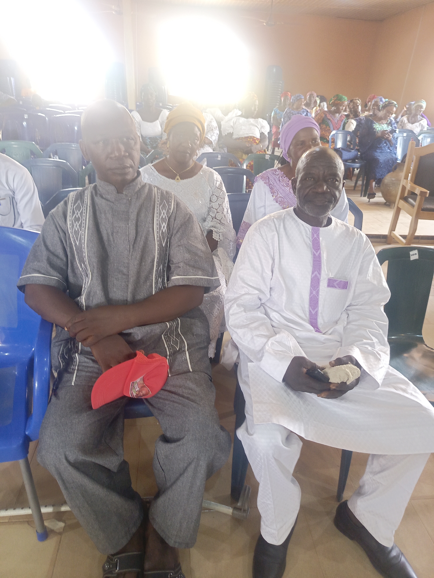 In Akwanga, Nigeria, the image shows two older men seated in the foreground of a room filled with seated women in the background. The man on the left wears a gray patterned outfit and holds a red cap. The man on the right is wearing a white outfit with a purple vertical strip and is holding a light-colored mass in his hands. Both men are seated on plastic chairs. Many women in the background wear head coverings and are seated in similar plastic chairs. The room is well-lit with a beige floor.