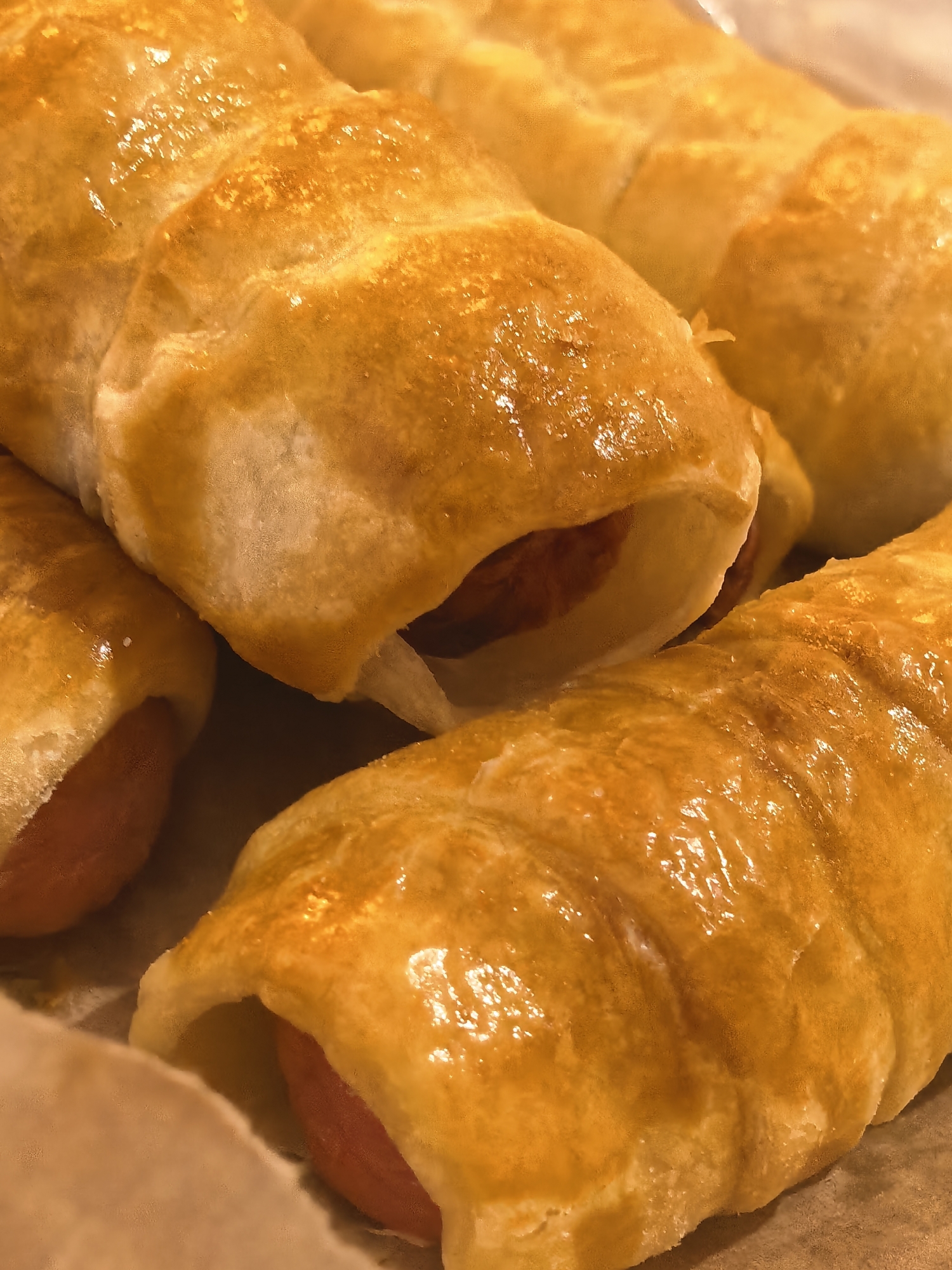 The media file is a close-up, overhead shot of several golden-brown pastry-wrapped sausages, commonly known as pigs in a blanket. The pastries are baked to a glossy finish, with visible layers and a slightly puffed texture. Some of the sausages are partially visible peeking out from the ends of the pastry. The lighting is warm, highlighting the rich color of the baked goods and creating reflections on their surfaces. The pastries are arranged in a way that suggests they have just been baked and are ready to be served. The background is a soft, out-of-focus brown, possibly indicating a baking sheet or parchment paper. There is no visible text or any indication of people, weather, or a specific time of day. The focus is entirely on the appetizing appearance of the food.