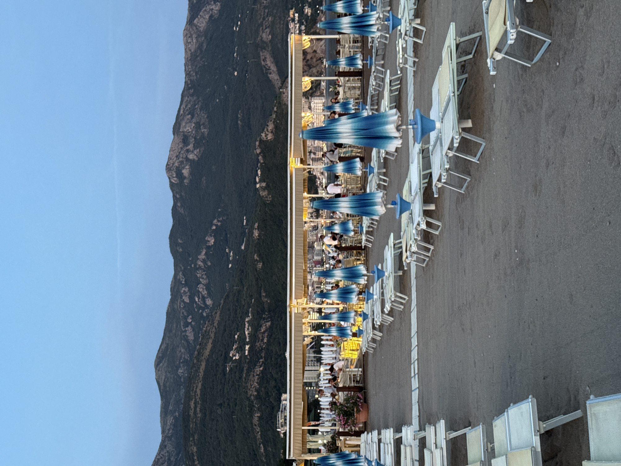 The image shows a beach resort in Boreti, Montenegro. The foreground is dominated by rows of empty lounge chairs and closed beach umbrellas with blue and white stripes. In the mid-ground, a covered outdoor restaurant or seating area is visible, with patrons seated at tables. The restaurant features white awnings and decorative lighting. Beyond the restaurant, more beach furniture and umbrellas are set up, and a body of water, likely the sea, is partially visible. The background consists of densely forested hills and mountains, with some buildings scattered on their slopes. The sky is a pale blue, suggesting either early morning or late evening light. The overall scene depicts a quiet resort area at a time when it is not fully utilized.