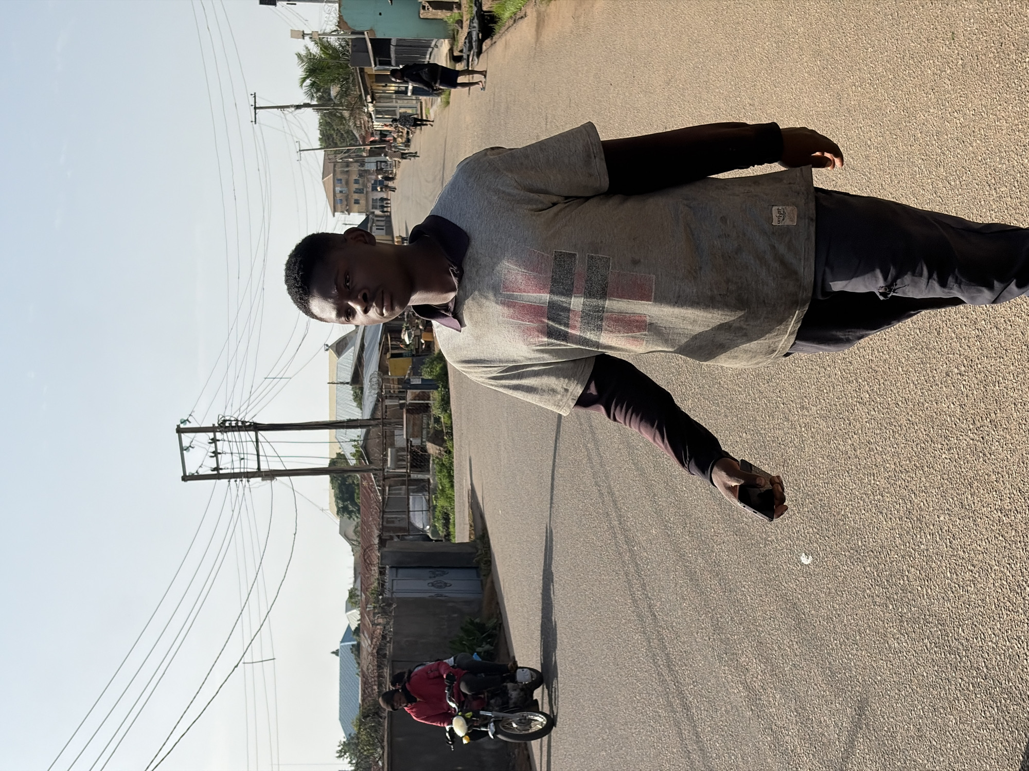 A young man stands on a paved road in what appears to be a residential area of Jos, Nigeria. He is wearing a gray t-shirt with a design on the front, dark long-sleeved shirt underneath, and dark pants. He is holding a black rectangular object in his right hand and looking directly at the camera.
In the background, there are houses, telephone poles with wires, and a person riding a motorcycle. Other individuals are visible further down the road. The sky is a clear pale blue. The scene is bathed in sunlight.