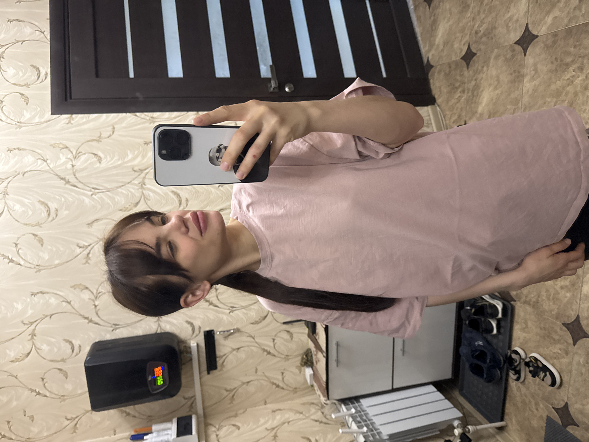 A woman with dark brown hair and a long ponytail is taking a selfie in what appears to be a hallway or entryway. She is wearing a light pink t-shirt and holding a smartphone with a light-colored case. The background features patterned wallpaper, a dark brown door with vertical stripes, and a tiled floor. Various household items are visible, including a black device with an orange digital display, a shoe rack with shoes, and a small white cabinet. There is no information available to conclude that this photo was taken in the city of Rostov-on-Don, Russia.