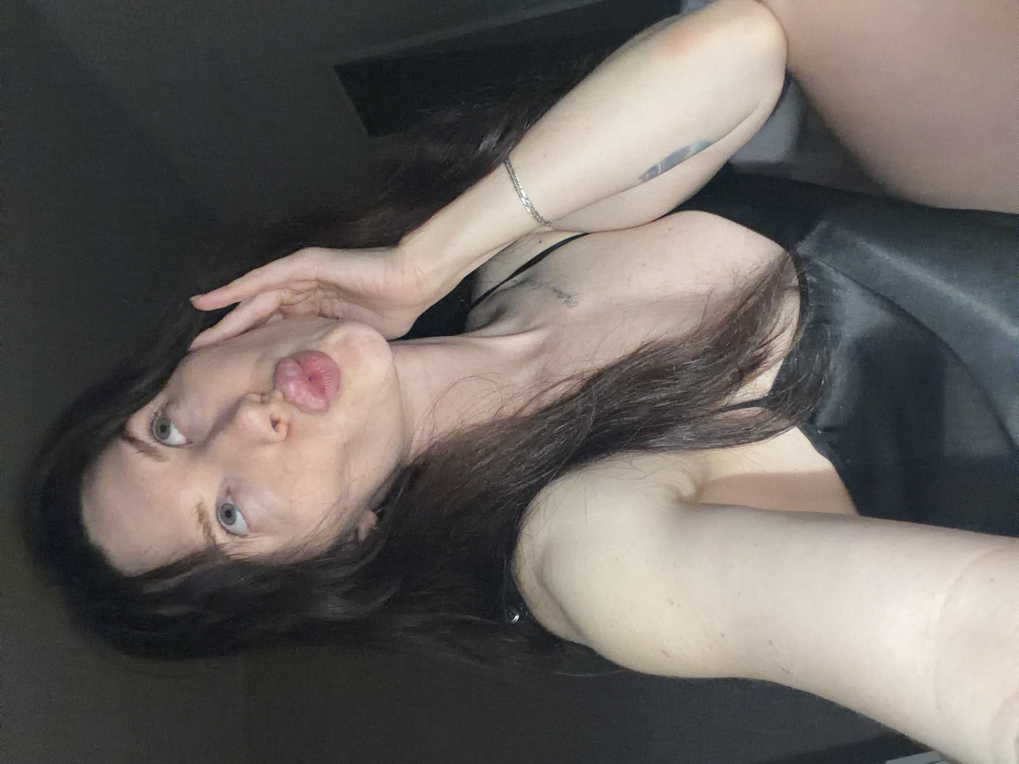 A woman is photographed from a low angle in a dark environment. She has long, dark hair, fair skin, and light-colored eyes. Her lips are pursed in a pouting expression. She's wearing a dark-colored garment that appears to be a satin top or dress. A silver bracelet is visible on her left wrist. There is a visible tattoo on her arm. The background is dark and undifferentiated. The location is unspecified and not discernable from the image.