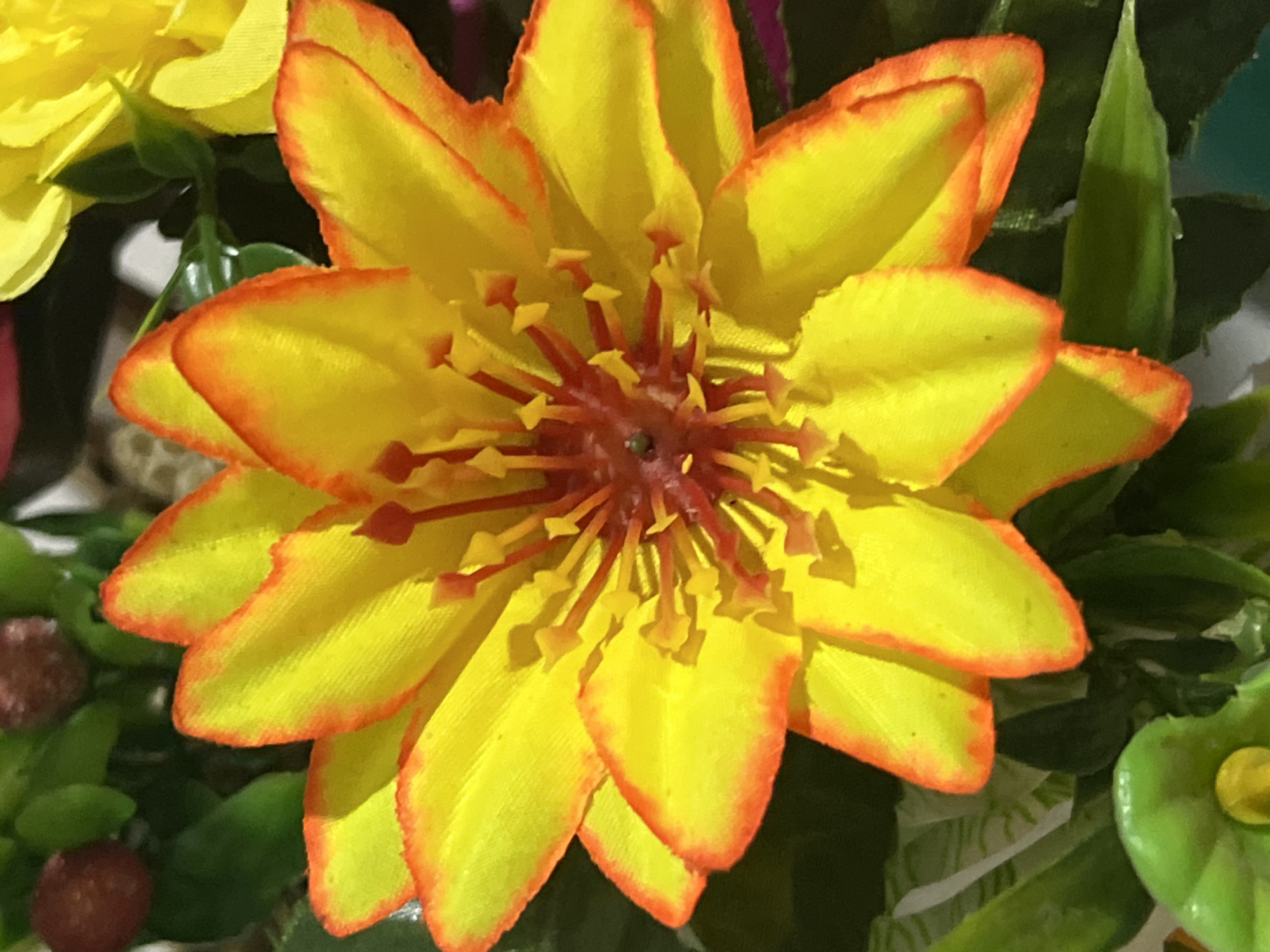 The image displays a close-up view of a vibrant yellow artificial flower with orange-red tipped petals. The flower has numerous red stamens tipped with yellow, clustered in the center. The background consists of blurred green foliage and what appear to be small red berries, suggesting a floral arrangement or decorative display. The lighting creates shadows within the flower's petals. The image does not contain any identifiable landmarks or features that would allow for pinpointing its location within Denpasar, Indonesia, or any other city.