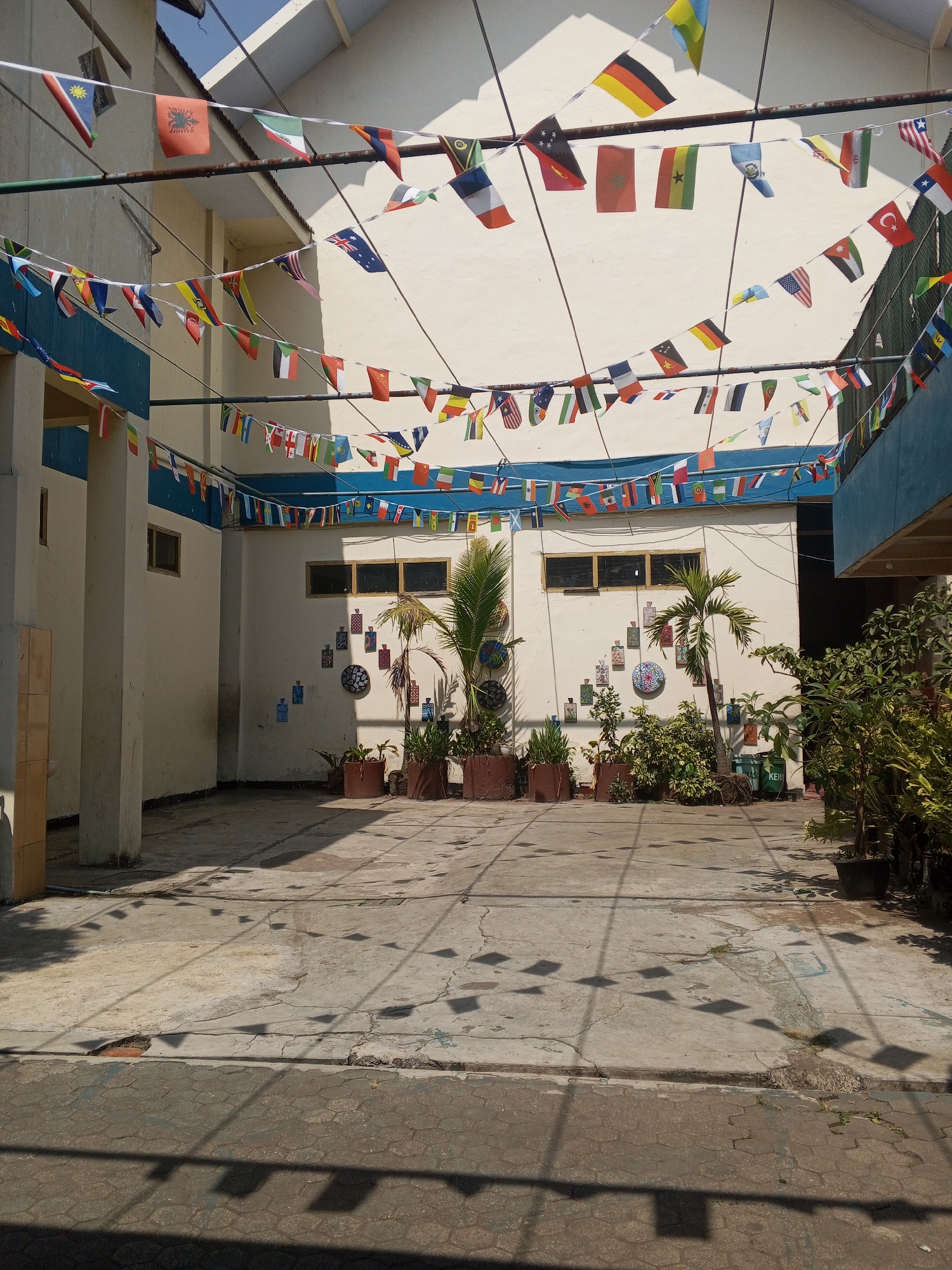 This image depicts an outdoor courtyard area, likely part of a school or community building, located in Turen, Indonesia. The courtyard is paved with concrete and is mostly empty, with shadows of flags and possibly overhead wires cast across the ground. The surrounding buildings are light-colored with blue trim along the upper sections. A prominent feature of the scene is the display of numerous small international flags strung across wires suspended above the courtyard. These flags are a mix of colors and designs, suggesting a celebration or educational display related to global diversity.
On the right side of the courtyard, several potted plants and small trees are arranged. One of these is a palm tree with visible roots. The wall of the building behind the plants is decorated with several square, colorful patterned tiles and a circular decorative element.
The lighting suggests it is daytime, with sunlight creating distinct shadows. The overall atmosphere is bright and somewhat open, with a sense of modest but cheerful decoration. There are no people visible in the image. The details about the location being in Turen, Indonesia, are external information not directly inferable from the visual content alone but are provided as context.