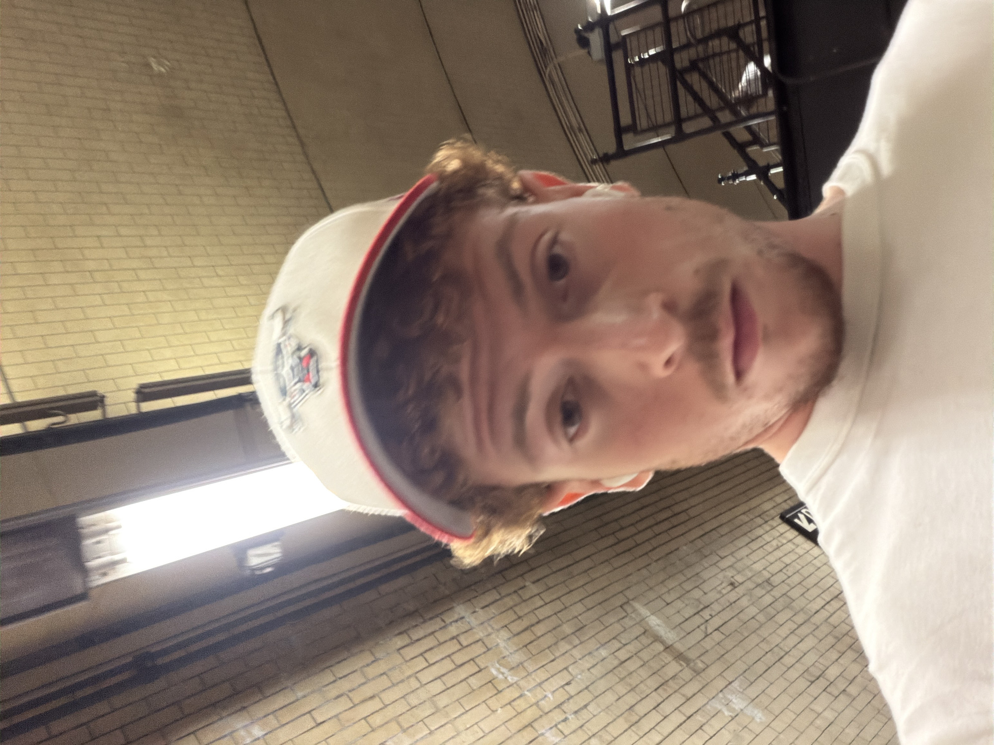 A man with light skin, light brown curly hair, and a beard stubble is shown in a selfie. He is wearing a white t-shirt and a baseball cap with a white brim and red trim. The background appears to be a New York City subway station with brick walls and fluorescent lighting. There is a black metal grate above. A black sign with white lettering is visible in the bottom right corner.