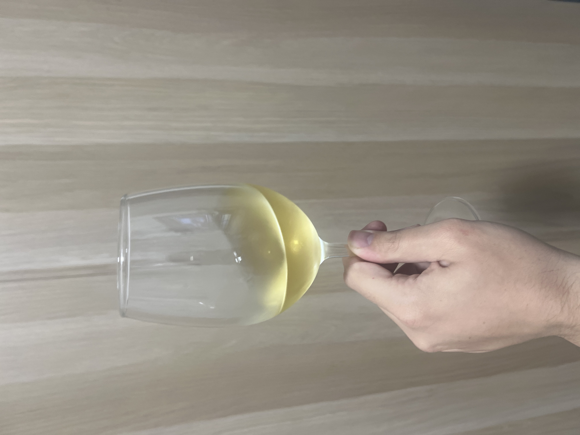 A wine glass containing a clear, pale yellow liquid is held horizontally by a light-skinned hand against a light brown wood-grain surface. The stem of the glass is positioned toward the hand's grasp. The location is not discernible from the provided image. There is no indication of the City of Mogán, Spain.