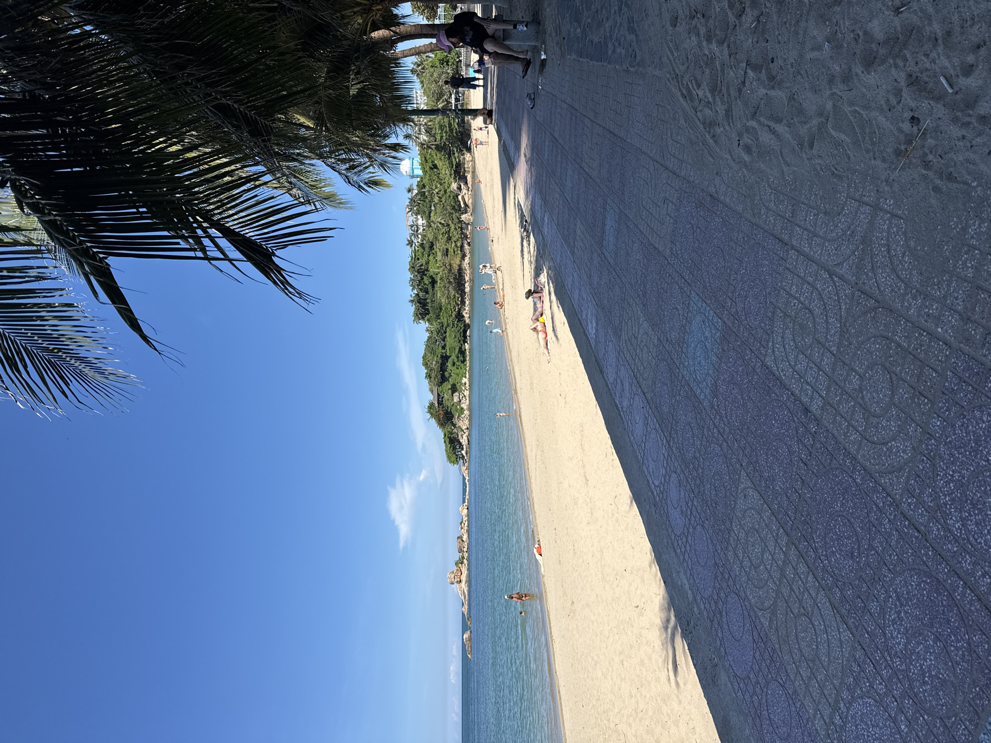 The image depicts a sunny day at a beach in Nha Trang, Vietnam. The foreground features a paved walkway with a patterned design, bordering a sandy beach. The beach itself is populated with several individuals engaged in various activities. Some people are in the shallow water, while others are sunbathing on the sand. The ocean is calm and blue, with a few rock formations and a distant tree-covered hill visible in the background. Palm trees line the edge of the walkway, casting shadows on the scene. The sky is clear and blue, indicating pleasant weather.