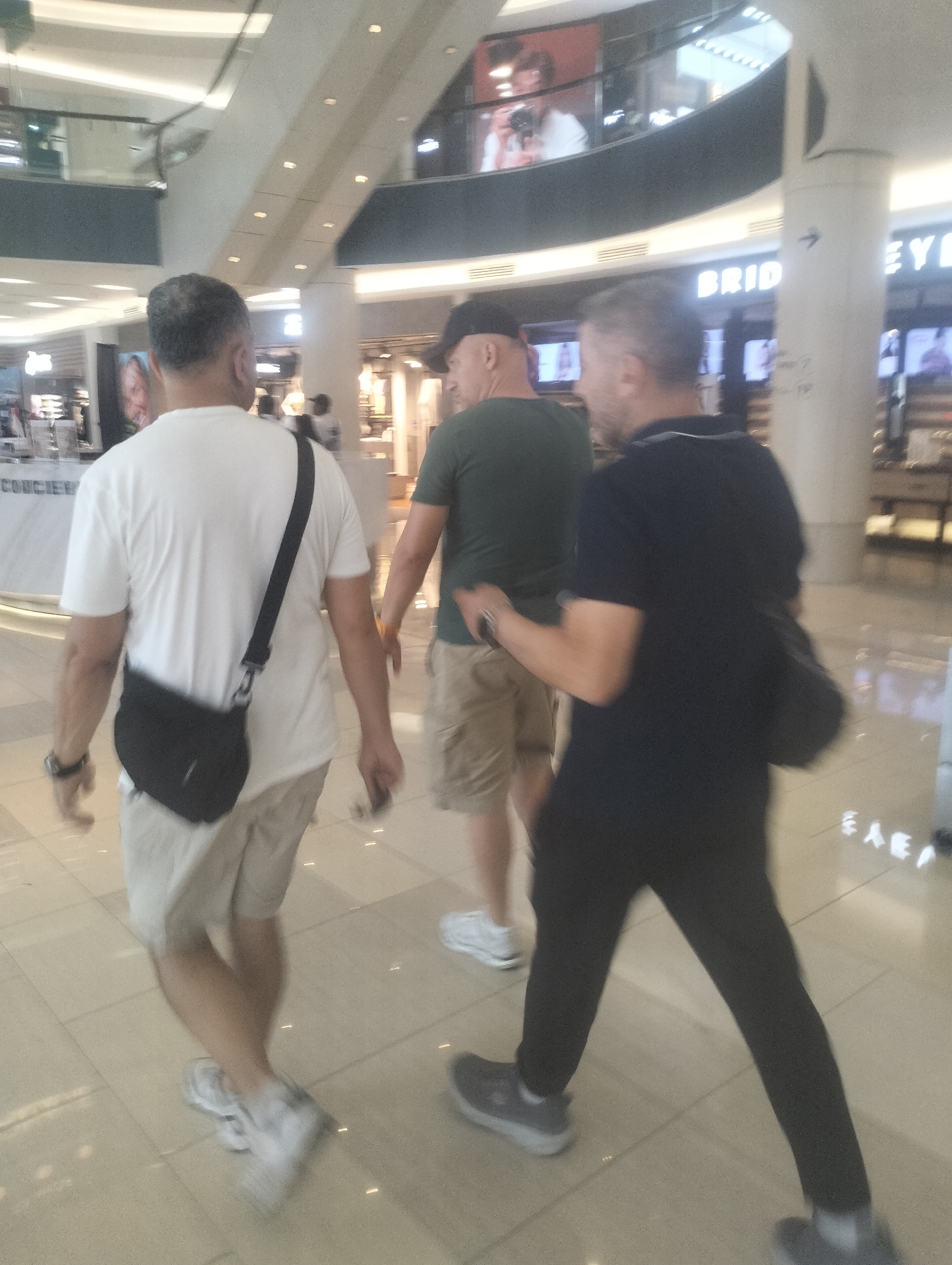 Three men are walking in a bright, modern indoor shopping mall. The man in the foreground on the left is wearing a white t-shirt and light-colored shorts, with a black crossbody bag. He has dark hair and appears to be middle-aged. In the center, another man is walking away from the camera, wearing a green t-shirt and tan shorts. He is bald and wearing a black baseball cap. To the right, a third man is walking in the same direction, wearing a dark blue polo shirt and dark pants. He has short, light-colored hair and is carrying a black bag over his shoulder. The mall environment features tiled flooring, large white pillars, and bright overhead lighting. In the background, elevated levels of the mall are visible, with advertisements and store displays. One visible advertisement in the background displays the word "BRIDGE". The style of the mall and the presence of specific signage suggests this location could be within a modern shopping complex in Surabaya, Indonesia, such as Tunjungan Plaza or Pakuwon Mall.