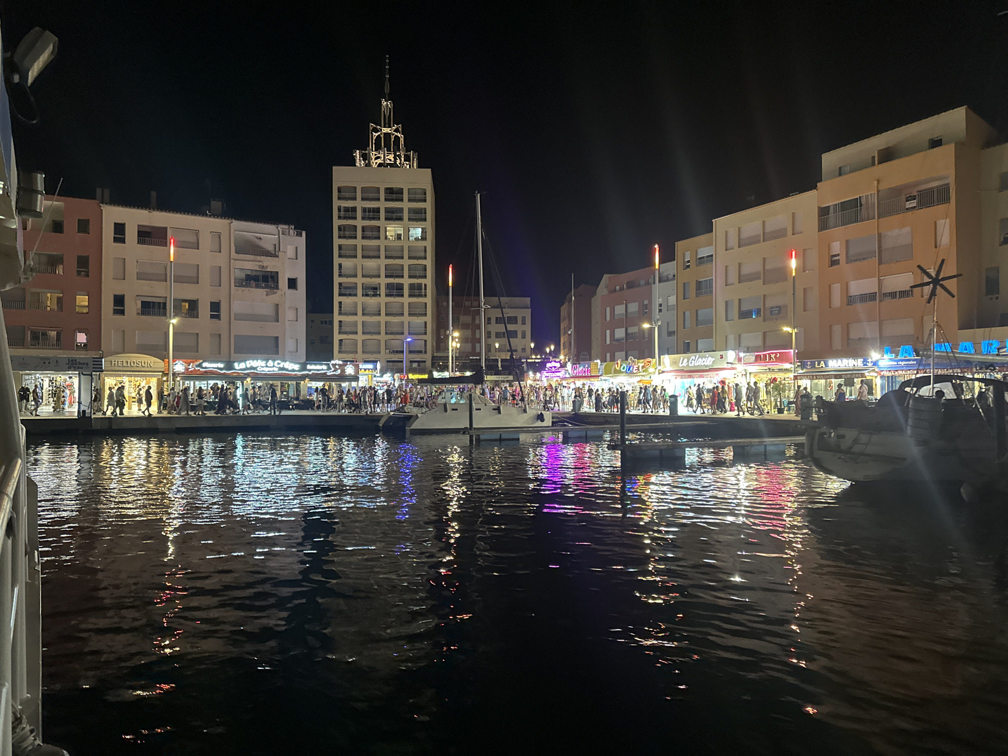 The image depicts a nighttime scene at a marina in Agde, France, likely the Grau d'Agde or Cap d'Agde. The waterfront is lined with buildings featuring shops and restaurants, illuminated by colorful lights. A prominent multi-story building with a distinctive antenna-like structure stands in the background. Several boats are docked in the water, reflecting the vibrant lights from the shore. People are visible walking along the promenade, suggesting a lively atmosphere. The water reflects the colorful illumination, creating a shimmering effect. The overall scene conveys a festive and bustling nighttime ambiance.