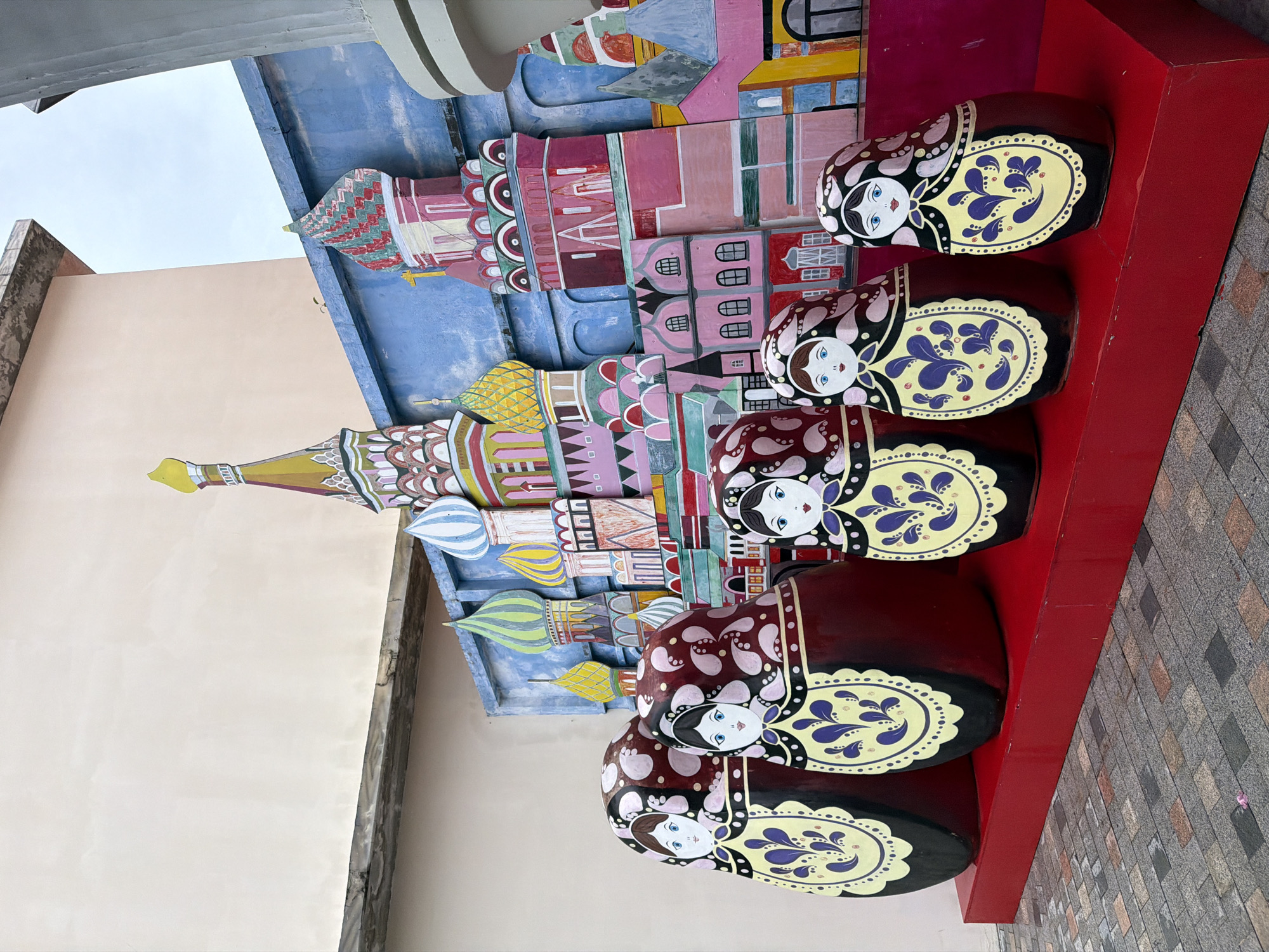 This image shows a street art display featuring large matryoshka dolls in front of a colorful backdrop depicting Russian architecture, likely St. Basil's Cathedral. The dolls are dark red with yellow and purple floral patterns and painted faces. The backdrop is a flat, multi-colored mural with various buildings and domes painted in bright hues. The scene is set outdoors on a paved walkway. This display is located at Sun World Danang Wonders in Da Nang, Vietnam, a location known for its theme park attractions and cultural replicas.