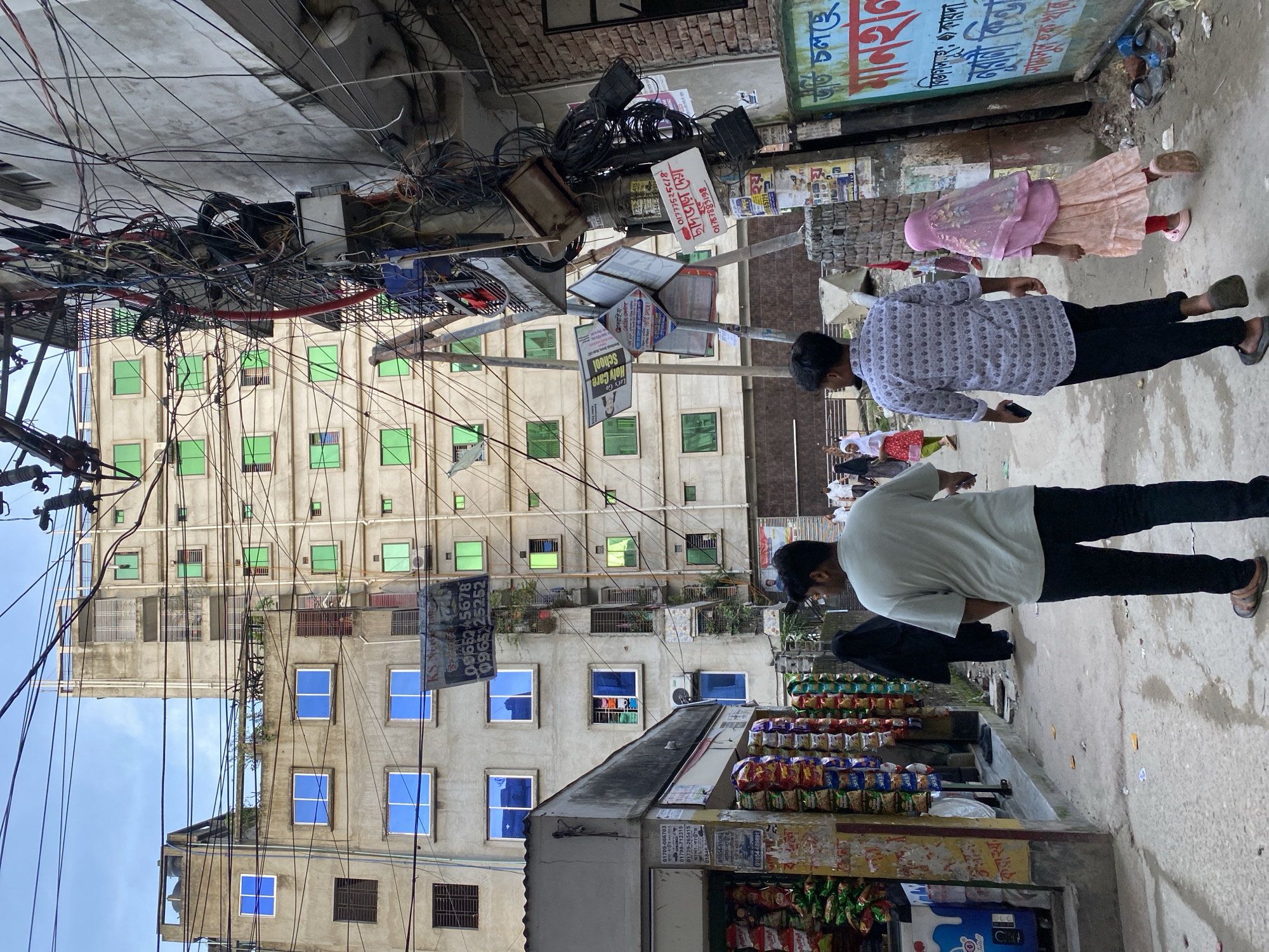 In a narrow urban street in Dhaka, Bangladesh, the scene is dominated by a dense network of electrical wires hanging precariously overhead. Buildings line both sides of the street, their facades a mix of concrete and brick, adorned with numerous windows, many featuring vibrant blue or green tinted glass. On the left, a small shop is visible, its shelves stacked with colorful bags of snacks. Several individuals are walking down the street. In the foreground, two men are walking away from the camera, one in a light-colored t-shirt and dark pants, the other in a patterned shirt and dark pants. A woman in a black burqa and a young girl in a pink hijab and dress are also walking away from the camera further down the street. The ground is paved and shows signs of wear, with some scattered litter. The overall atmosphere is one of a busy, densely populated city environment.