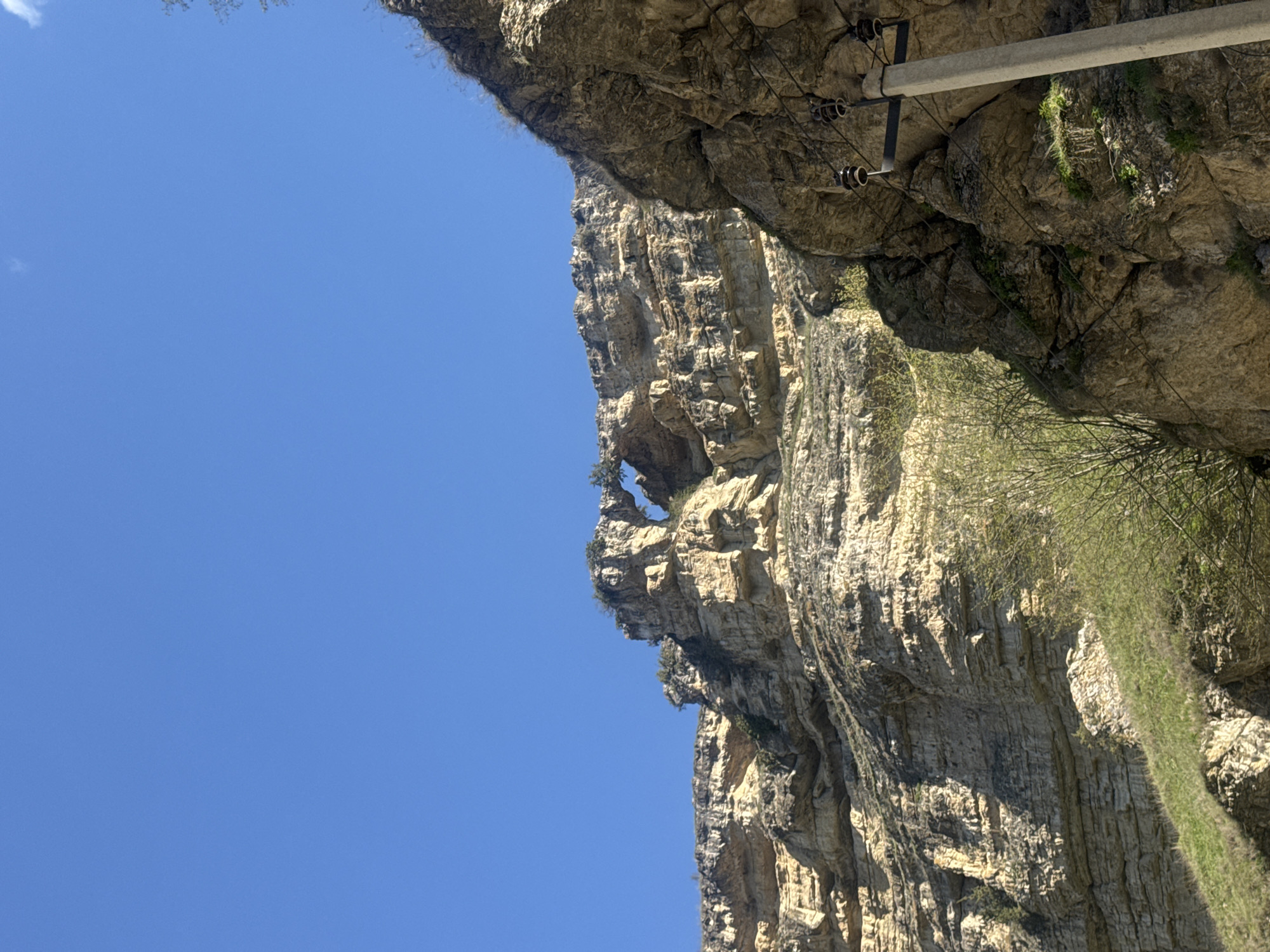 A vertical, low-angle shot shows a massive rock formation under a clear blue sky. The rock face is layered and textured with shades of brown and gray, featuring a natural arch near the top. Sparse vegetation clings to the rock, and a concrete structure with electrical wires extends along the top right. This image is taken at Kyondelen, Russia.