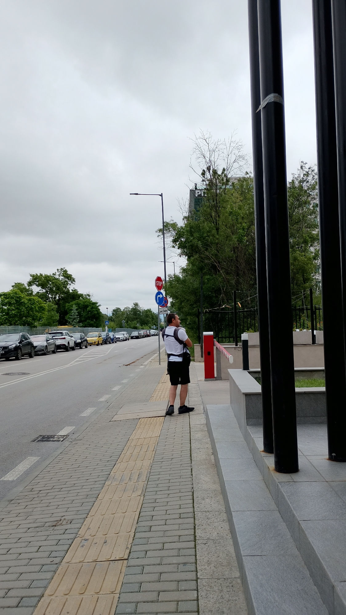 A man stands on a sidewalk next to a street with parked cars. He is wearing a white t-shirt, black shorts, and a backpack. He is looking away from the camera and holding a walking stick. The sidewalk is made of gray and yellow paving stones. There are traffic signs on the street, including a stop sign and a directional sign. The street is lined with trees and buildings in the background. The sky is cloudy.