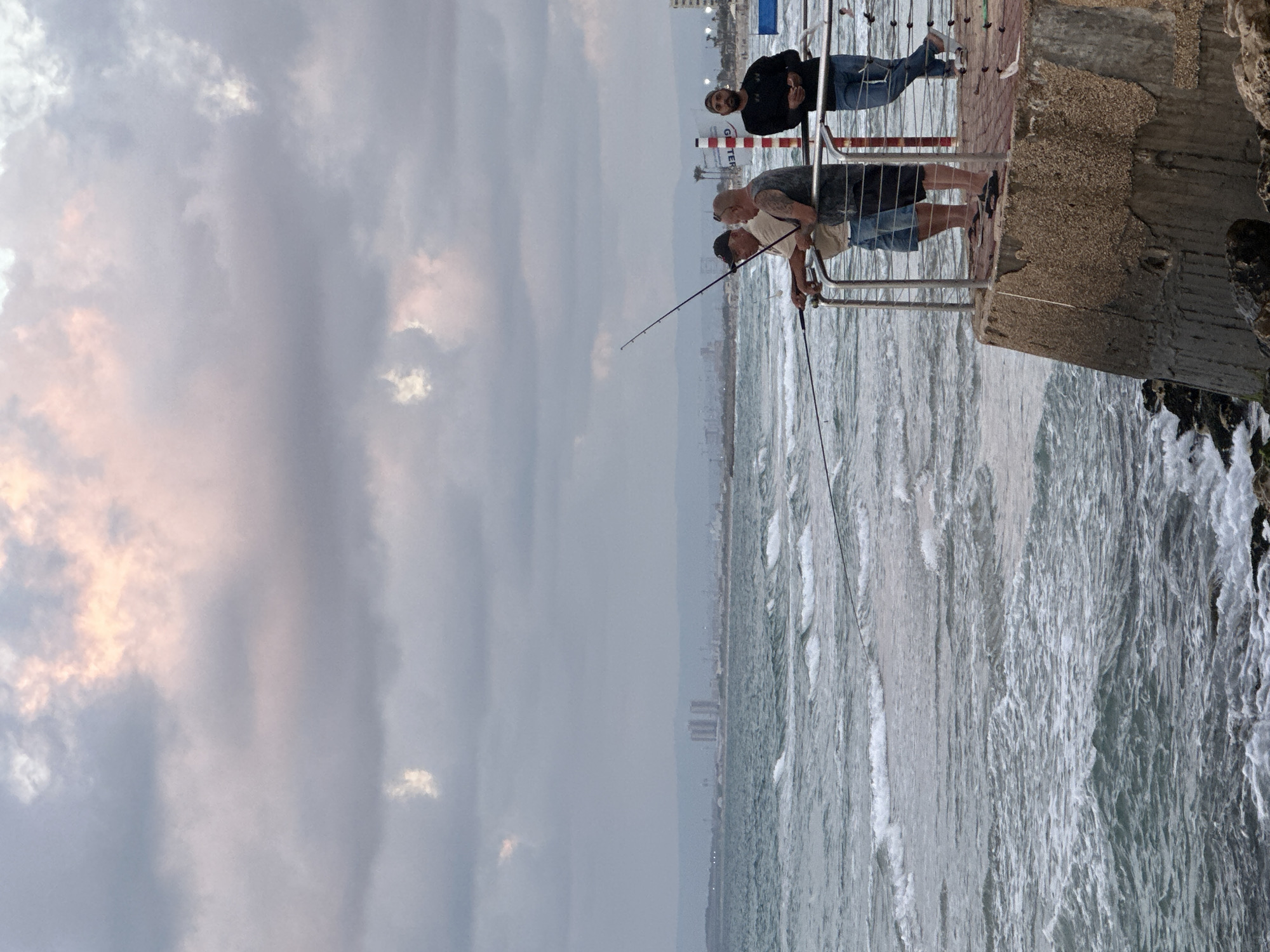 Four men are on a pier, fishing in the sea. Three of the men are standing together near the railing, with two of them holding fishing rods. The fourth man stands further to the right, looking out at the water. The sky is cloudy, with some pink and orange hues from the setting sun visible. In the distance, a city skyline can be seen across the water. The pier is made of concrete and has a metal railing. The water is choppy, with waves crashing against the shore. This scene is likely the Haifa Bay area of Haifa, Israel, given the urban skyline and coastal setting.