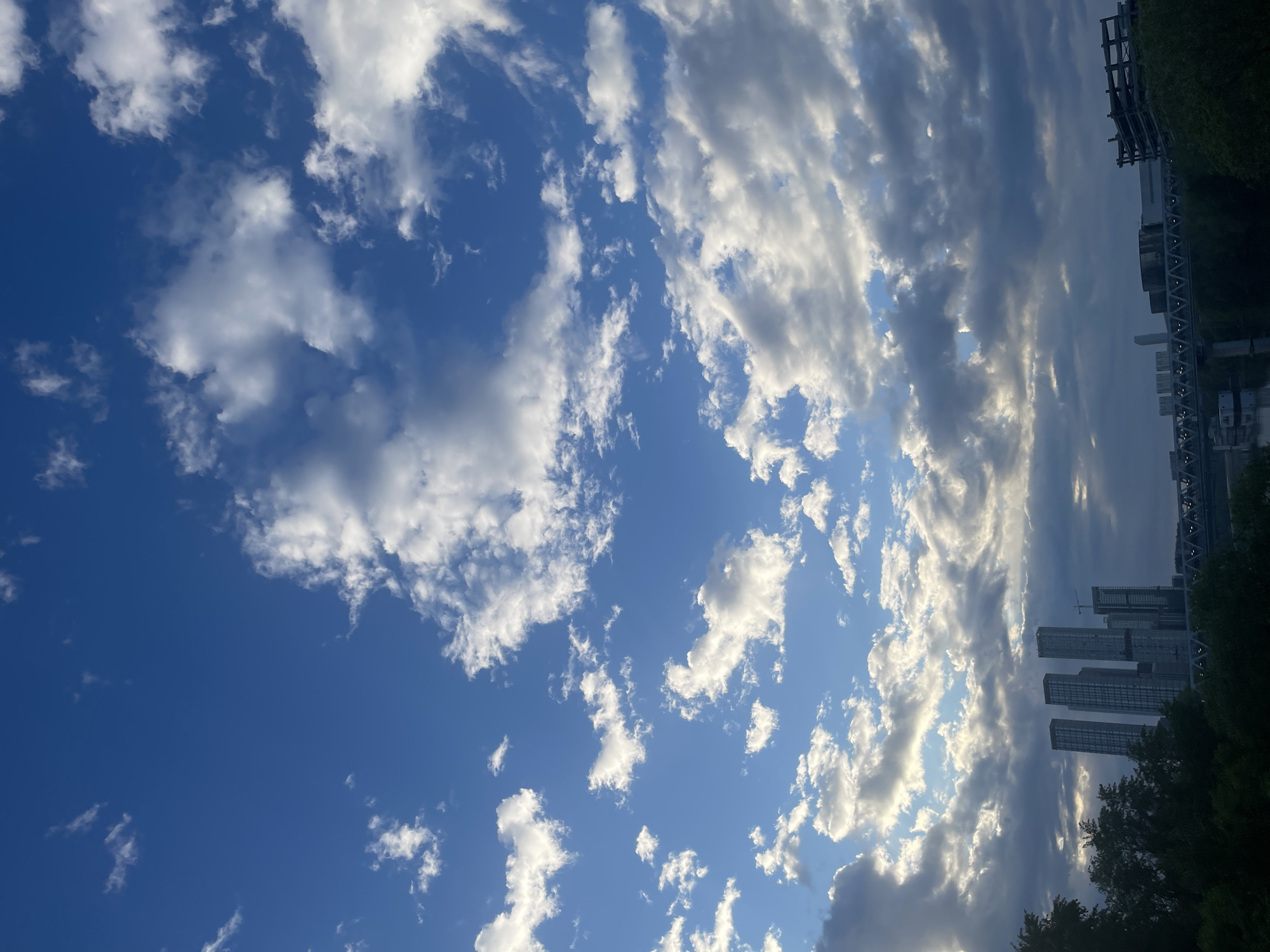 The image shows a daytime view of the sky above Khimki, Russia. The sky is primarily blue with scattered white clouds. The clouds are more concentrated on the right side of the image, where they partially obscure the skyline of the city. Several tall buildings are visible among the clouds, with foliage from trees below the buildings. A structural framework, possibly a bridge or overpass, is also visible at the top right. The image is rotated slightly, as the buildings appear angled relative to the horizontal.