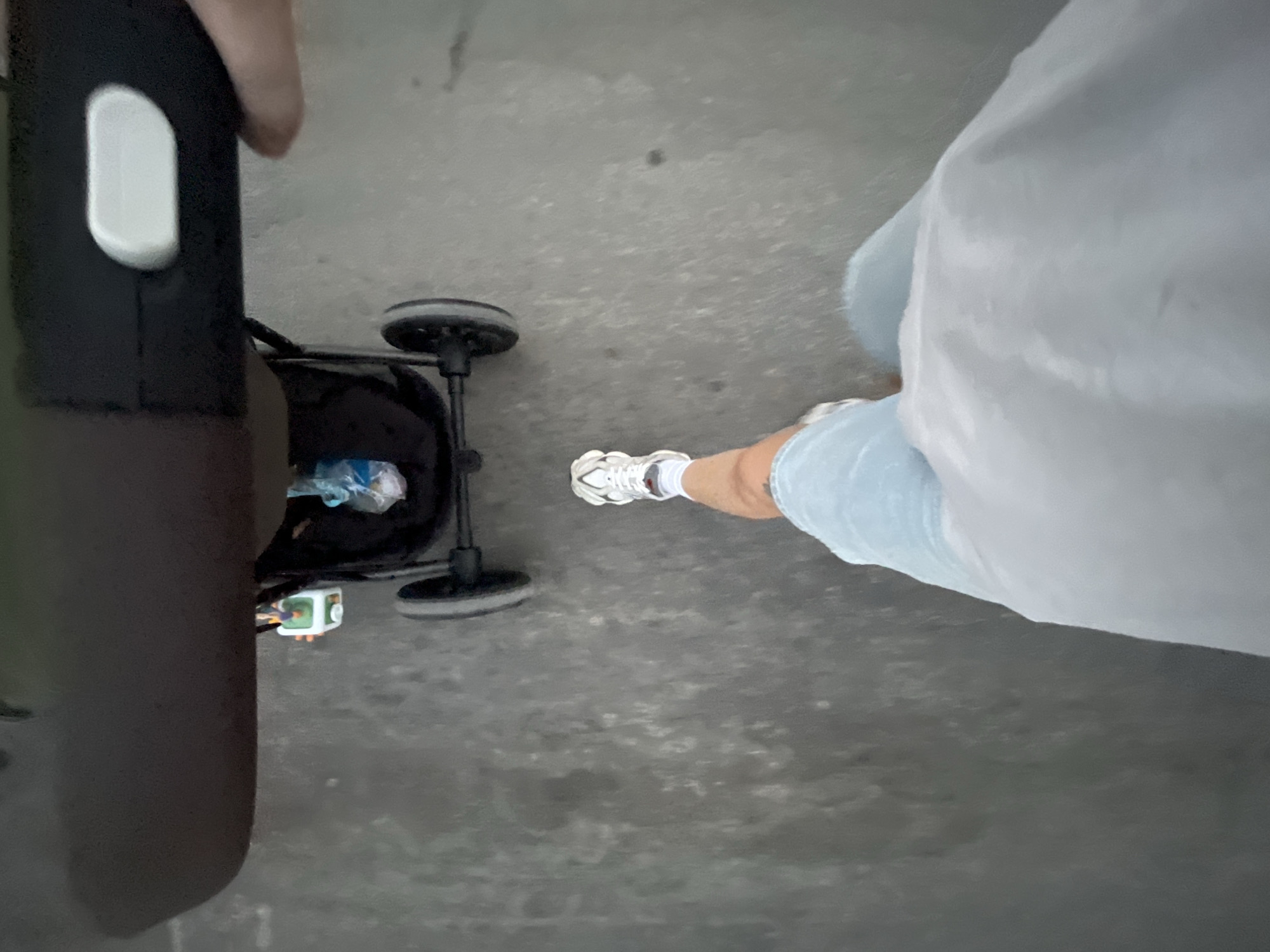 The image is a first-person perspective shot of a person pushing a stroller down a paved street, likely in Limassol, Cyprus. The stroller is black with a brown handle and a white button or lever on the handle. A toy is attached to the stroller. The person is wearing light blue shorts, white socks, and white athletic shoes. The ground is a textured, gray pavement. The lighting suggests it is daytime.