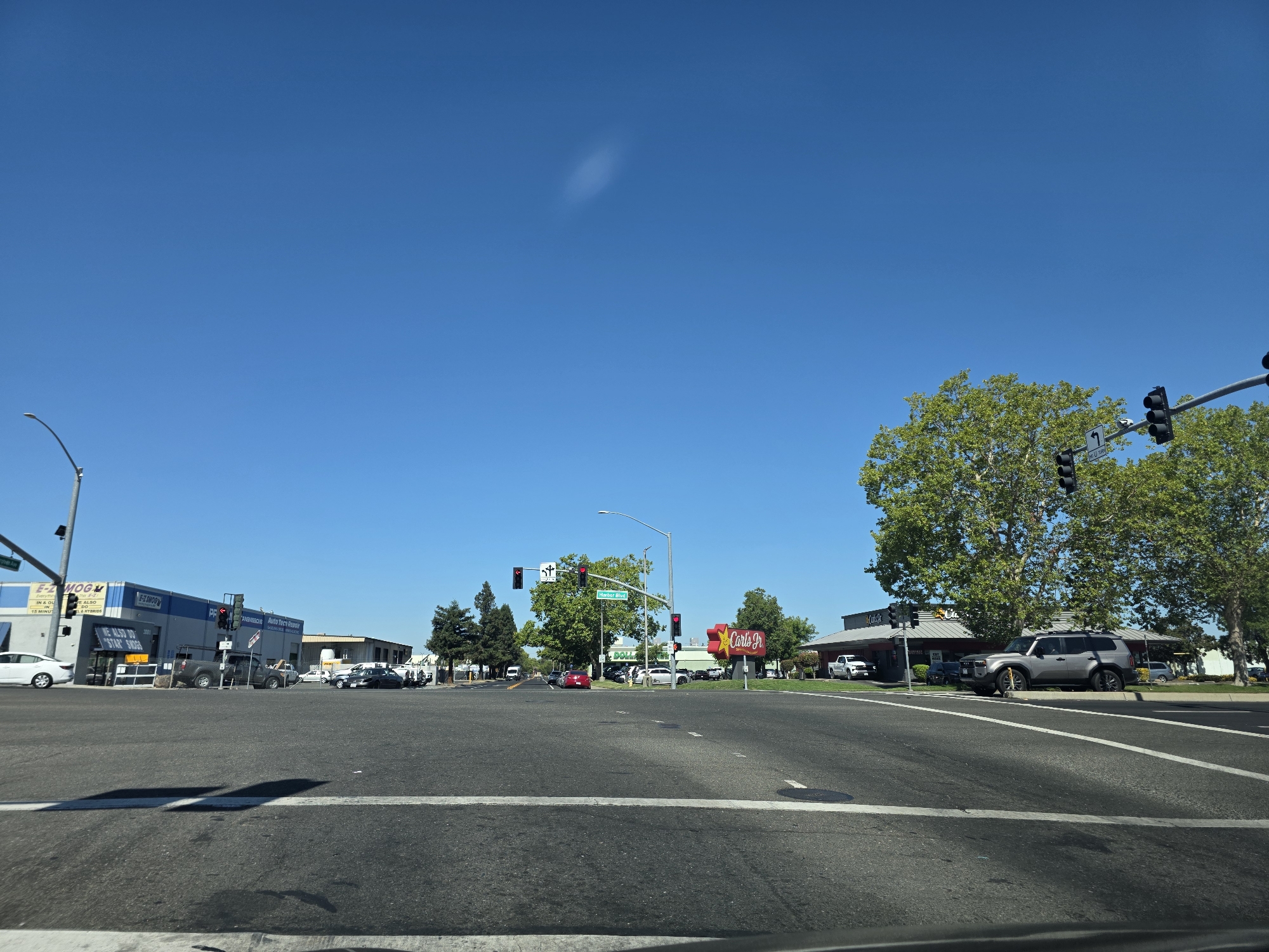 This image captures a wide-angle view of a street intersection on a clear, sunny day. The sky is a vibrant, cloudless blue, suggesting a warm, pleasant atmosphere. The foreground is dominated by asphalt, with white lane markings indicating a multi-lane intersection.
On the left side of the road, a commercial building is visible with signage that includes "E-Z SMOOG" and "IN & OU VE ALSO LA HYBRIDS". Next to it, another building displays "ANSMISSIONS Auto Tecy Repor" and "WE ALSO DO 'STAR' SWOGS". Several cars are parked and driving in this area.
In the center of the intersection, traffic lights are visible, with one showing a red light. Street signs indicate "Martin Blvd". A red car is stopped at the intersection.
On the right side of the intersection, a prominent "Carls Jr." restaurant sign with its distinctive star logo is clearly visible. Another building, possibly associated with the restaurant, is also present. A grey SUV is parked in front of it, and several other vehicles are in the background. Large, leafy trees provide shade and add greenery to the scene. The overall setting is a typical suburban commercial area in the United States, likely during daylight hours due to the bright sunshine and strong shadows. There are no people clearly visible in the image.