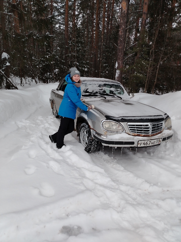 The image depicts a snowy, forested outdoor scene in or near Chaikovsky, Perm Krai, Russia. A young female, dressed in a grey knit beanie, a light blue hooded winter jacket, a patterned scarf, black pants, and black winter boots, stands on the driver's side of a silver sedan. She is smiling towards the camera, with her right hand resting on the snow-covered hood of the vehicle. The sedan, possibly a GAZ Volga Siber, is heavily covered in a fresh layer of snow, with multiple icicles hanging from its front bumper. The front grille shows visible rust, and the front left wheel, partially submerged in snow, is fitted with what appear to be snow chains or similar traction devices. A Russian license plate, "Y 467 HE 59," is clearly displayed on the front bumper. The vehicle is positioned on a snow-covered track or unpaved road, surrounded by deep snow drifts. A dense coniferous forest, with snow-laden trees, forms the background under an overcast sky.
