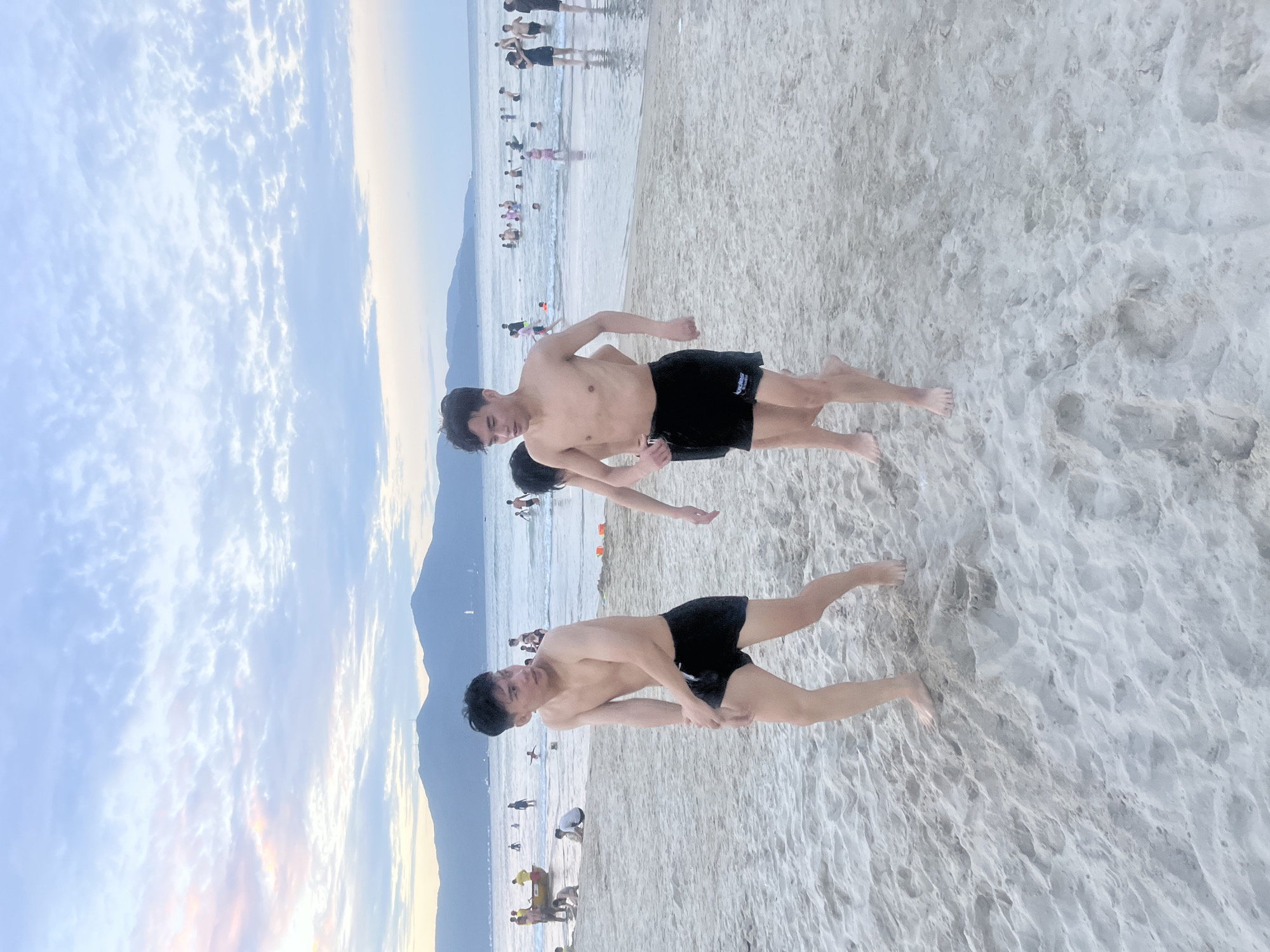 This image captures a group of young men on a beach in Da Nang, Vietnam, likely during the late afternoon given the soft lighting and overcast sky. The scene is set against a backdrop of the sea and a distant mountainous coastline. In the foreground, three young men are walking on the sandy beach, all wearing dark swim trunks. The man on the left is looking to his right, with his arms bent at the elbows. The man in the center is slightly behind and to the right of the first man, with his head turned and his arms extended. The third man is mostly obscured, visible from the chest down, walking to the right. In the middle ground, the sea is populated with numerous people swimming and wading. The far background shows the hazy outline of mountains. The beach is covered in fine, light-colored sand with some ripples and footprints. The sky above is a pale blue with scattered, wispy clouds tinged with soft colors, suggesting either sunrise or sunset.