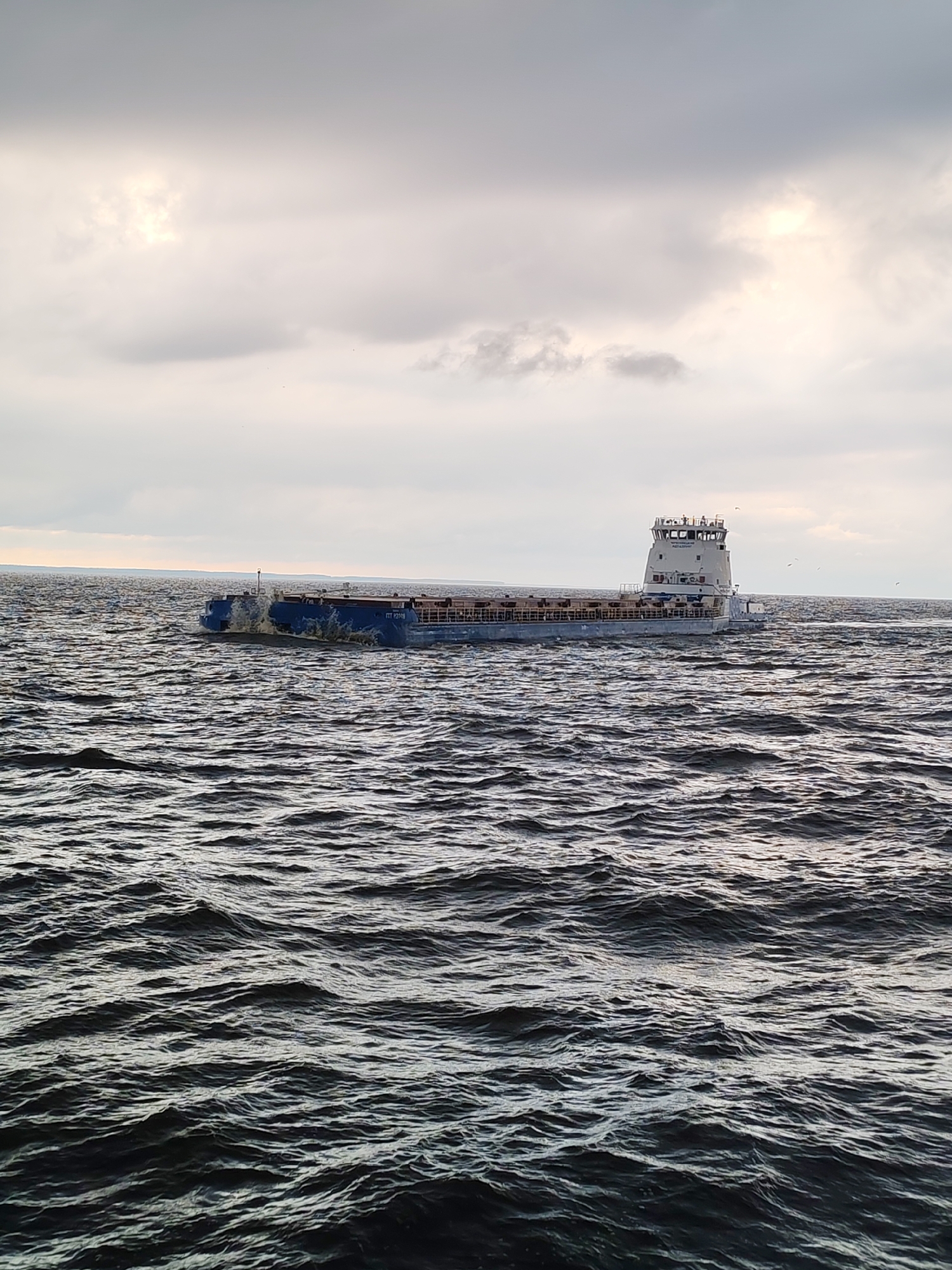 A cargo barge moves across choppy water under an overcast sky. The barge is long and flat with a raised superstructure at the stern, containing windows and what appears to be a navigation bridge. The water is a deep, dark blue with numerous waves, suggesting a windy or rough condition. The sky is filled with gray clouds, with some patches of brighter light indicating the sun may be breaking through or is low on the horizon. There are a few small, dark shapes in the sky that could be birds. The barge has some visible text on its side. The scene suggests a utilitarian setting, likely a body of water used for shipping or transport. The weather appears cool and possibly blustery, giving the overall scene a somewhat dramatic or somber mood. The time of day is unclear due to the overcast conditions, but the light suggests it could be morning or late afternoon.