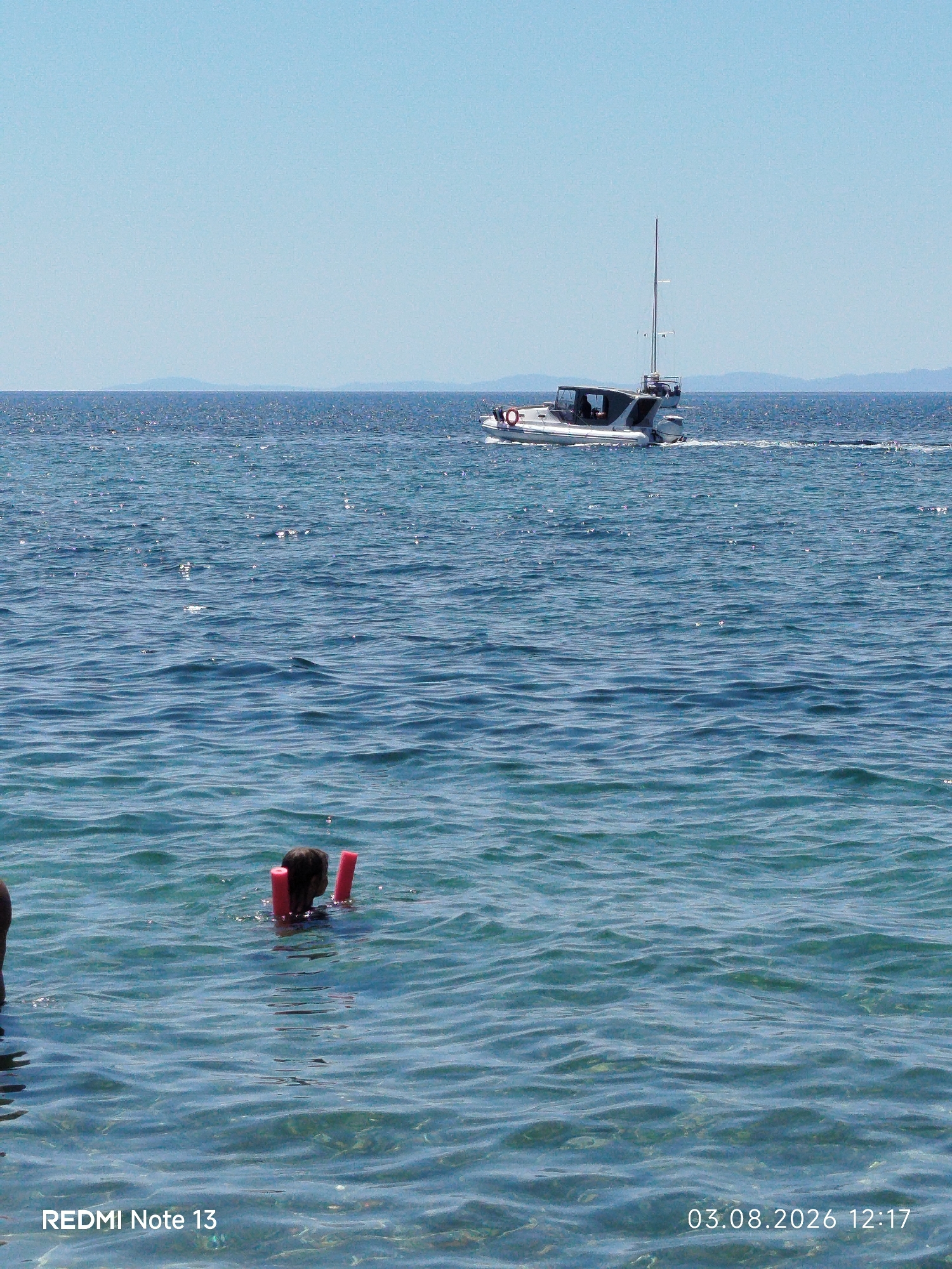In the foreground, a person is swimming in clear blue water, holding onto two red floatation aids. The water is slightly choppy with sunlight glinting off the surface. In the middle ground, a white motorboat with a mast is cruising across the sea, leaving a wake behind it. The background shows a hazy, blue sky meeting the distant, dark blue horizon, with faint outlines of landmasses. The image is dated "03.08.2026 12:17" and has the watermark "REDMI Note 13". The scene is consistent with a coastal location in Sithonia Municipal Unit, Greece, known for its clear waters and sailing activities.