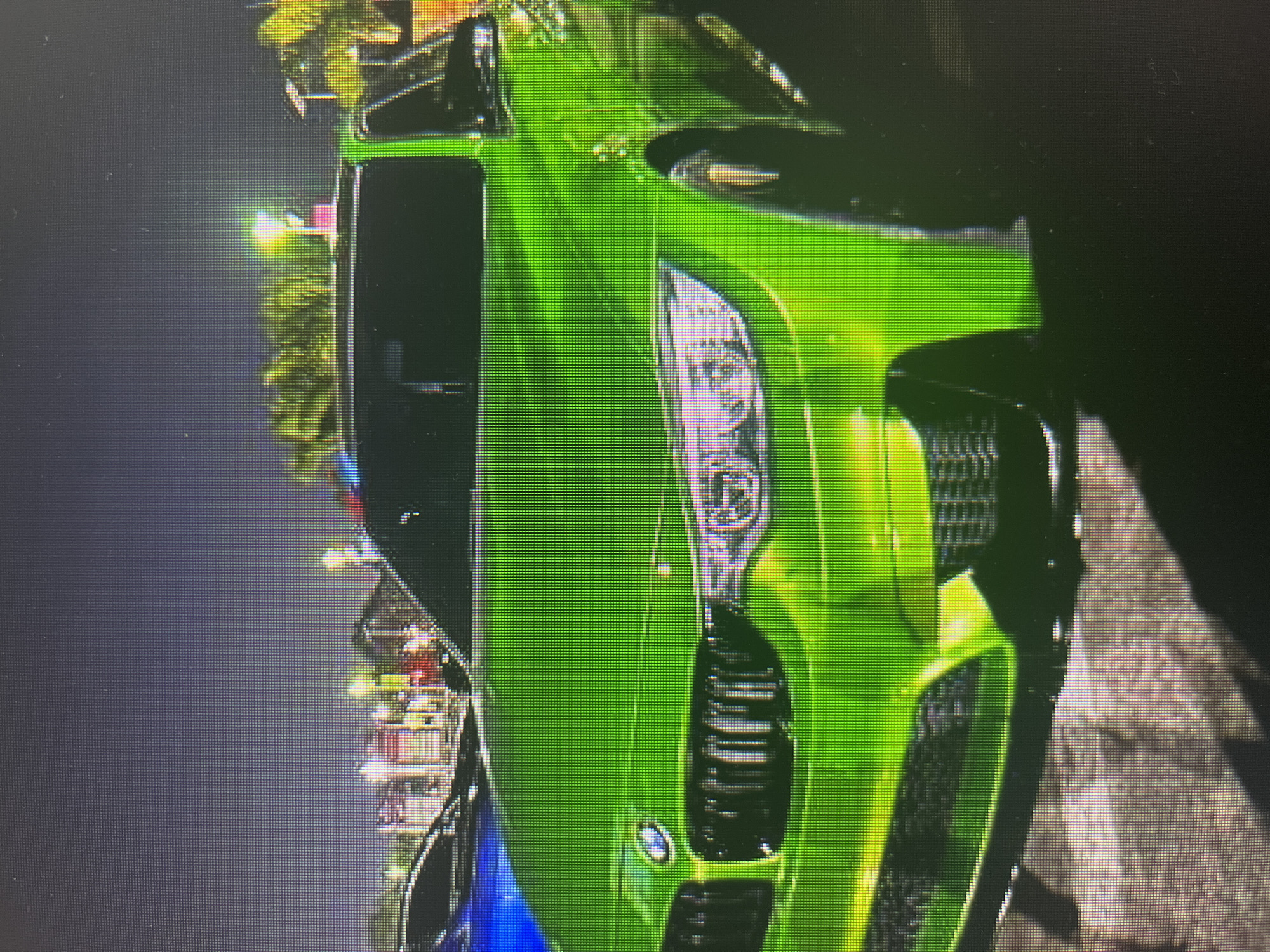 The image shows a bright green BMW M4 parked outdoors at night. The car is the primary subject, with its front end clearly visible. It has a distinctive grille with vertical slats and sharp headlights. The car is illuminated by external light sources, creating reflections on its glossy surface. In the background, other vehicles, including a blue car, are partially visible. There are also trees and buildings in the distance, with some lights from what appears to be a commercial or entertainment area. The ground in the foreground is paved. The scene appears to be in Varna, Bulgaria, possibly at a car meet or a public parking area.
