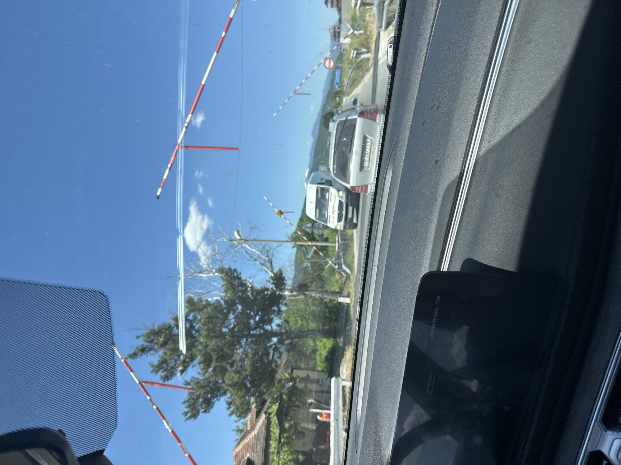 The image, taken from inside a vehicle, shows a railroad crossing in Unknown, Unknown. The railroad crossing gate is down, marked with red and white stripes. Two white vans are visible beyond the crossing. On the dashboard of the vehicle, there's a display showing "2026/05/29 Friday 11:45" and "Seat ventilation." Outside, the sky is blue with some clouds. A tree and a building are also visible.